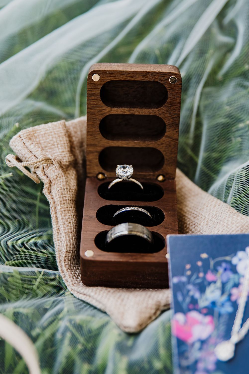 Elegant wooden ring box with diamond engagement ring and wedding bands on burlap pouch, surrounded by greenery.