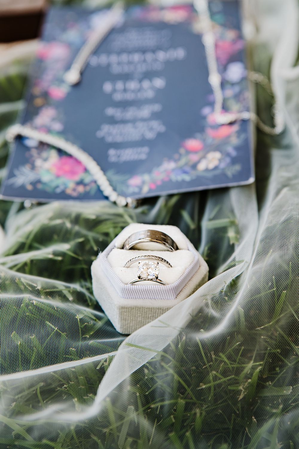 Wedding rings in a velvet box beside a navy floral invitation on sheer fabric and green grass.