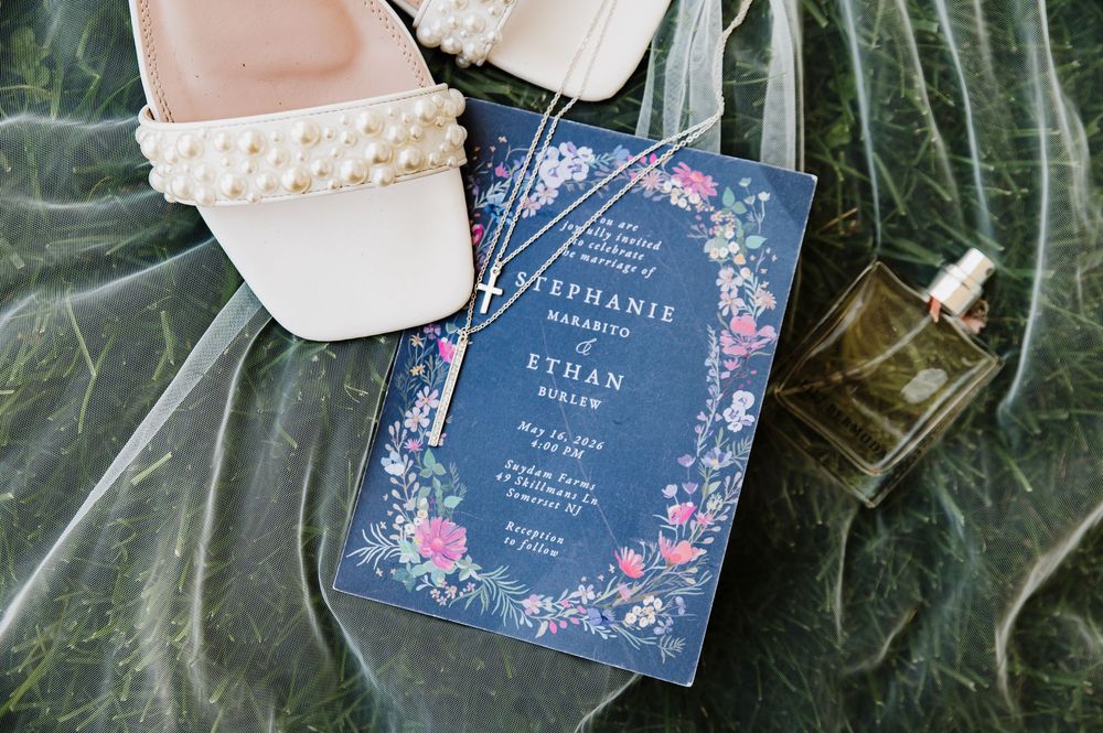 Navy blue floral wedding invitation with pink flowers beside pearl heels and perfume bottle on green leaves.