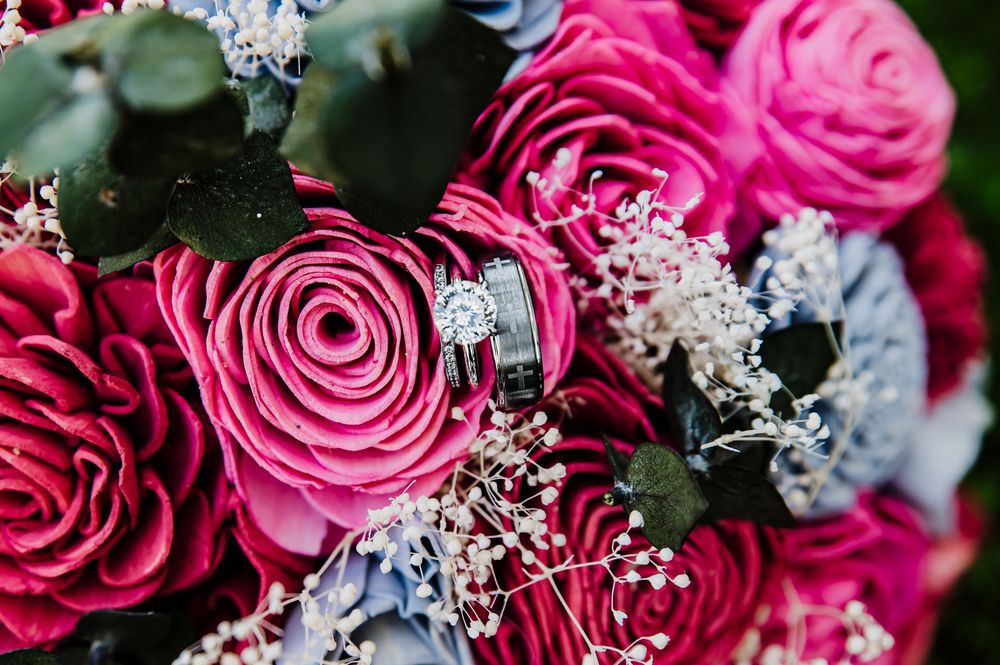 Wedding rings resting on vibrant pink and red roses in a bridal bouquet with blue flowers and greenery.