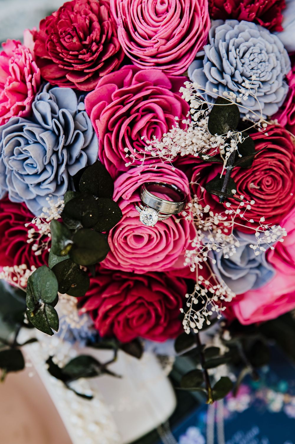 A vibrant wedding bouquet of pink and blue roses with baby's breath, adorned with two silver wedding rings.