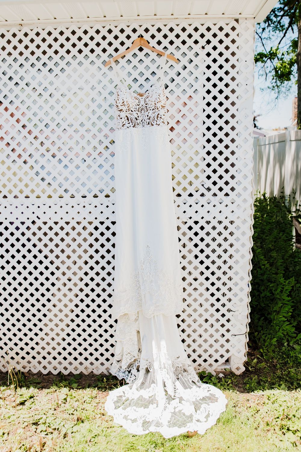 Elegant white wedding dress with lace train hanging on a wooden hanger against a white lattice fence outdoors.