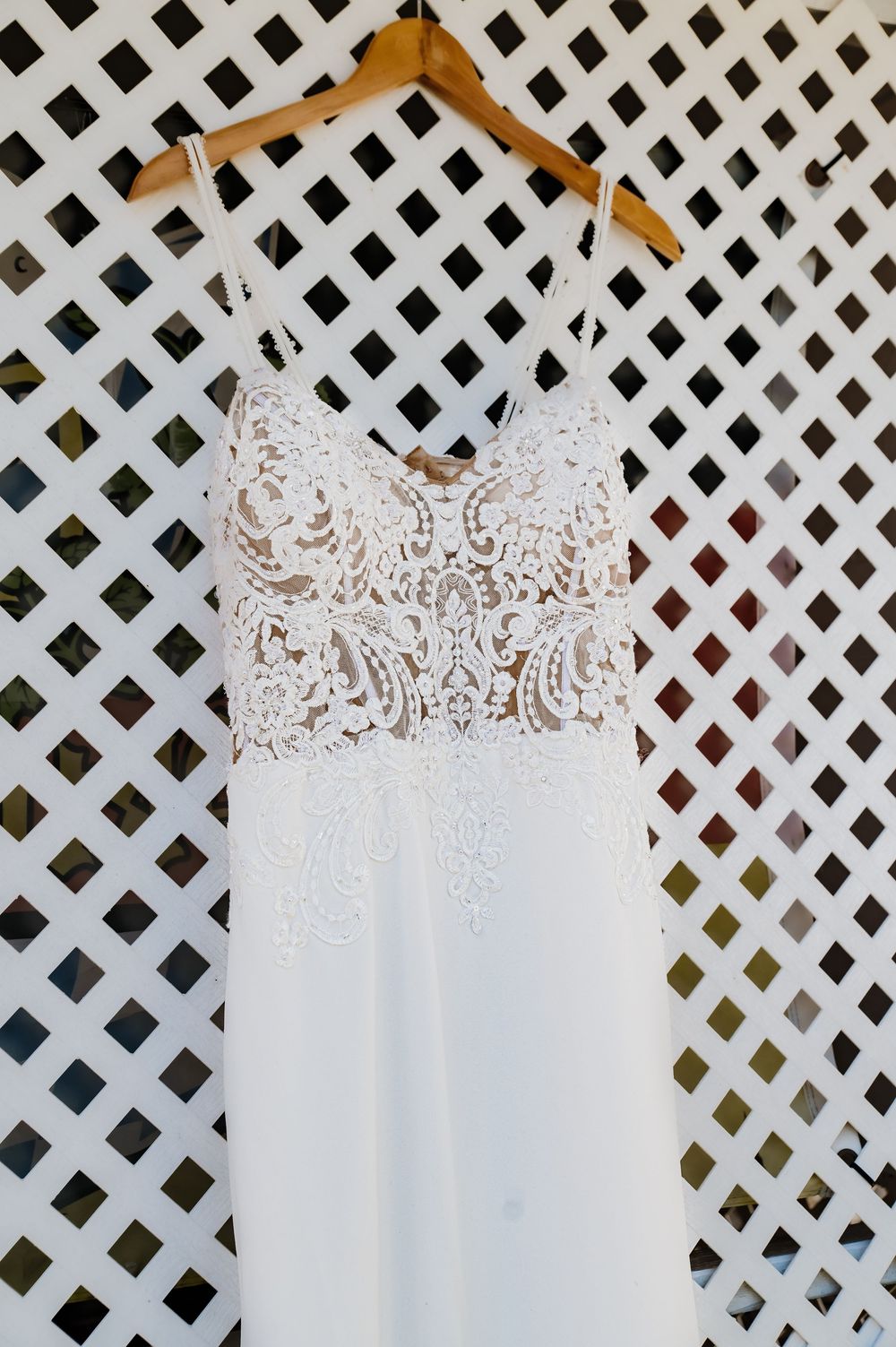 White lace wedding dress with spaghetti straps hanging on a wooden hanger against a white lattice background.