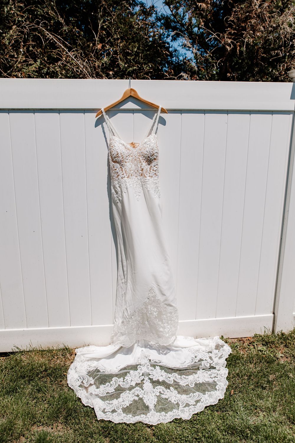 Elegant white lace wedding gown with train hanging on wooden hanger against white fence outdoors.