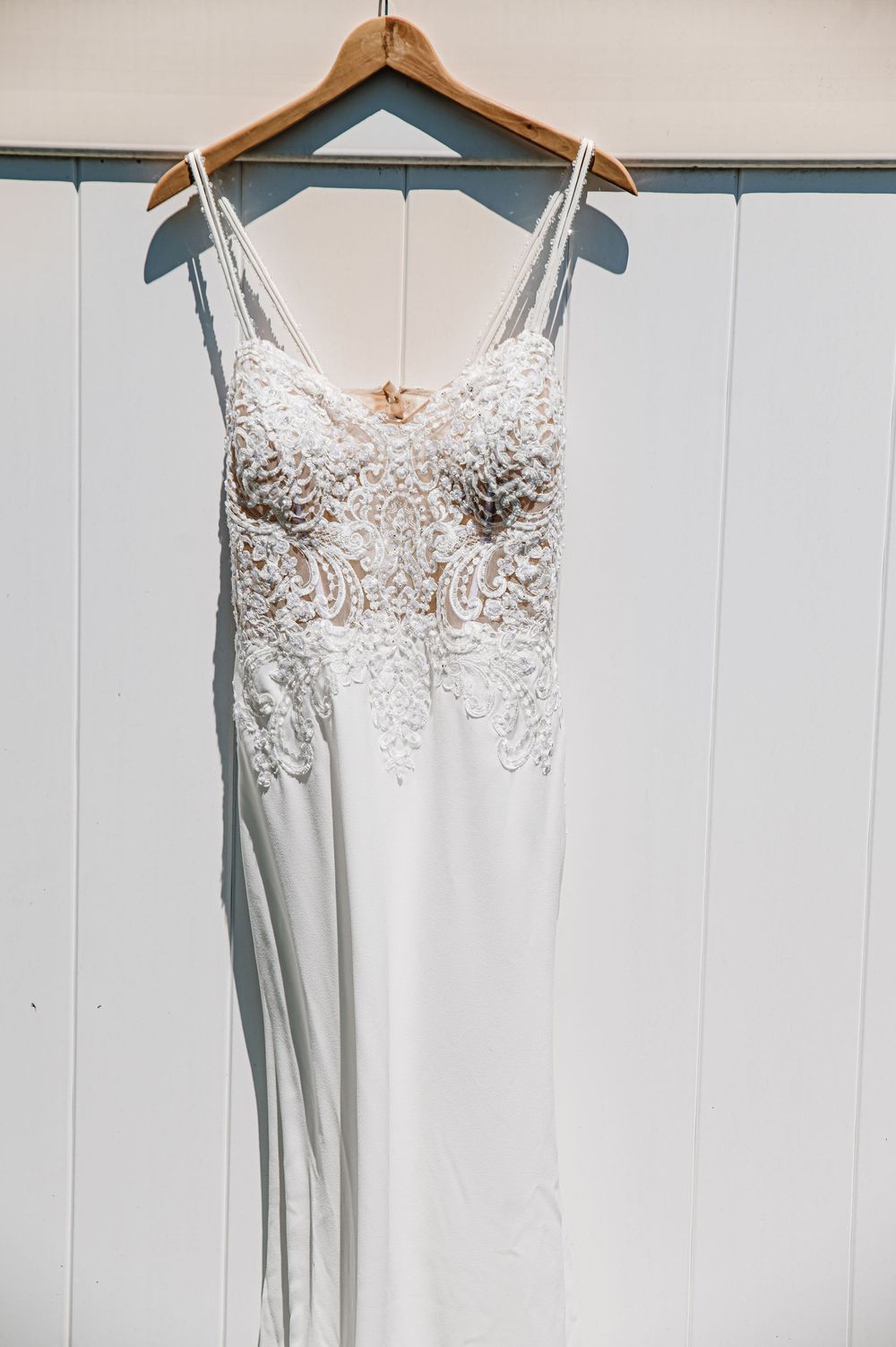 Elegant white beaded wedding gown with spaghetti straps hanging on a wooden hanger against a white paneled wall.