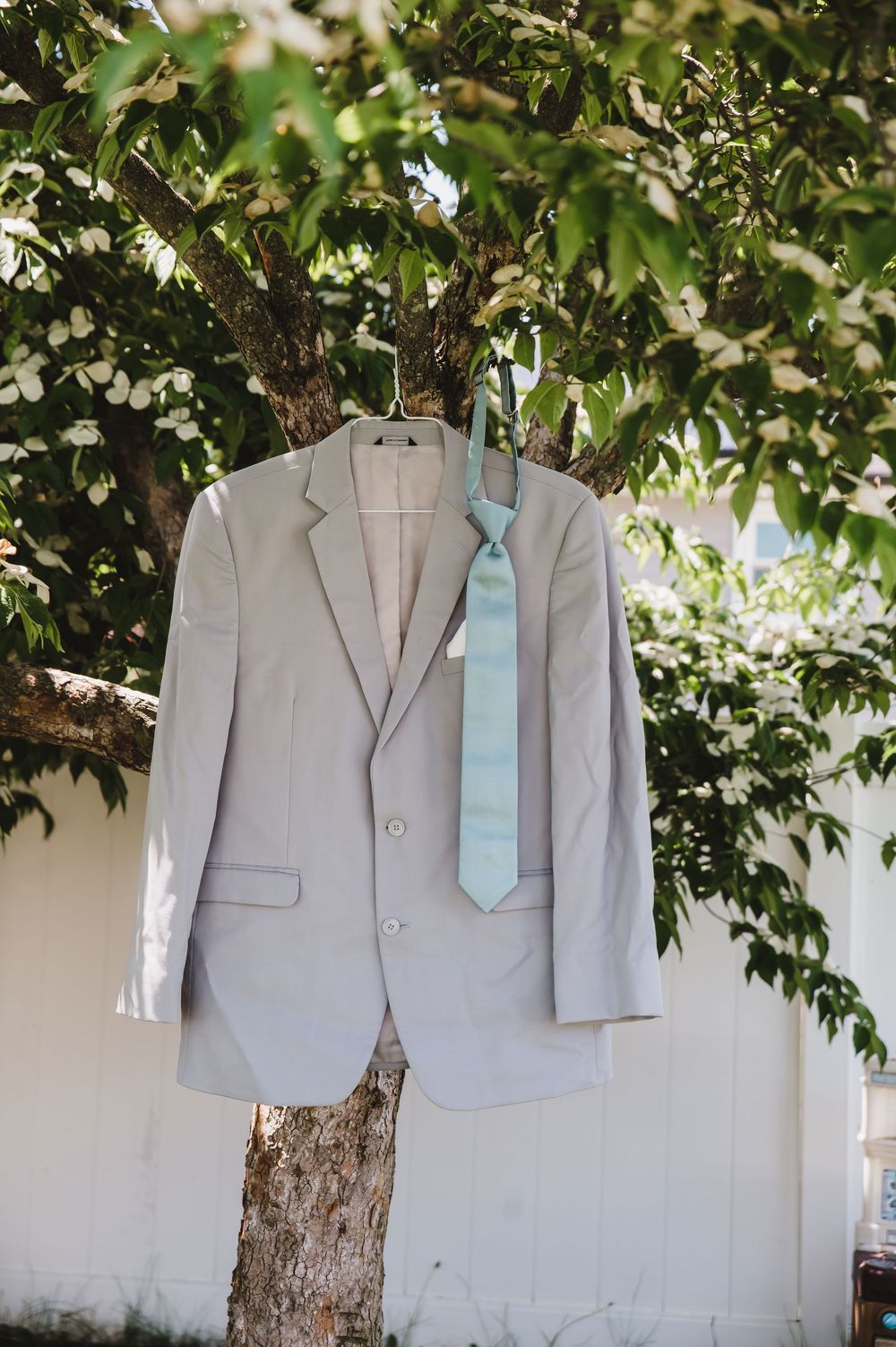 Gray suit jacket with light blue tie hanging on a tree branch outdoors at a wedding venue.