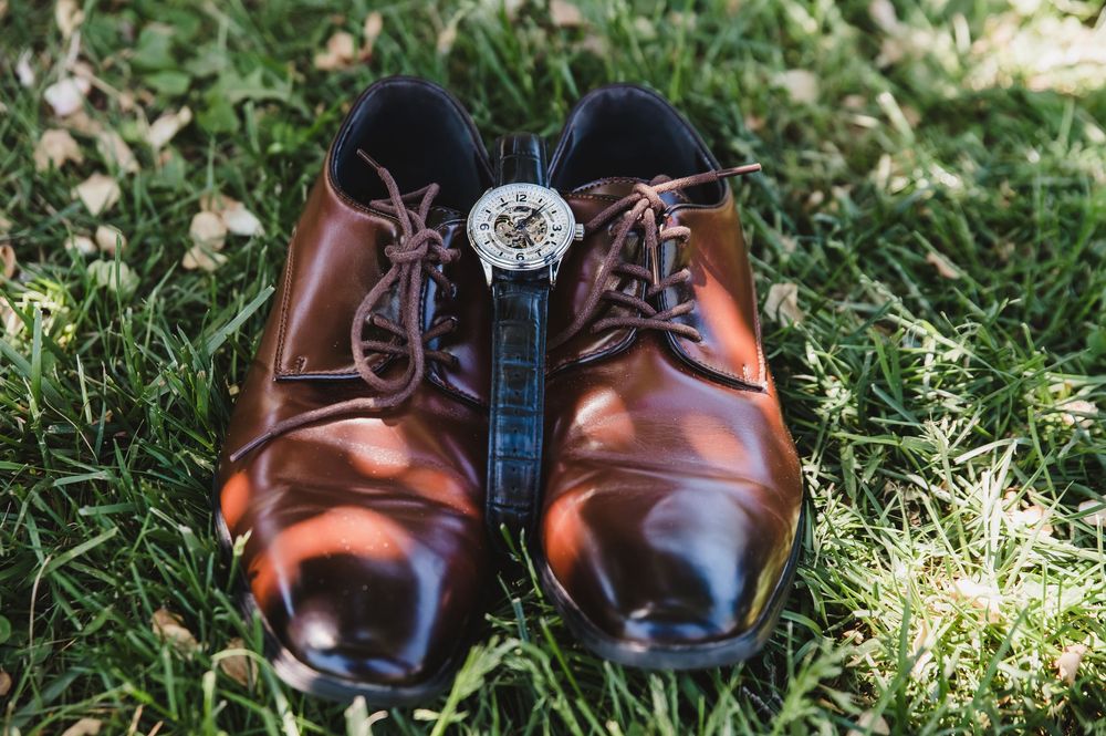 Brown leather dress shoes with laces on green grass, accented by a silver mechanical watch placed between them.