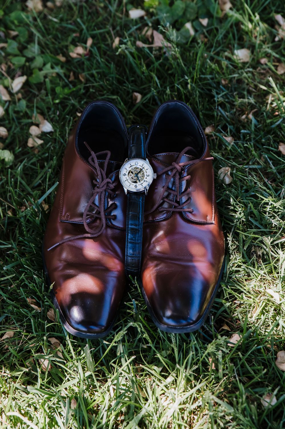 Polished brown leather dress shoes with laces and a decorative watch placed between them on green grass.