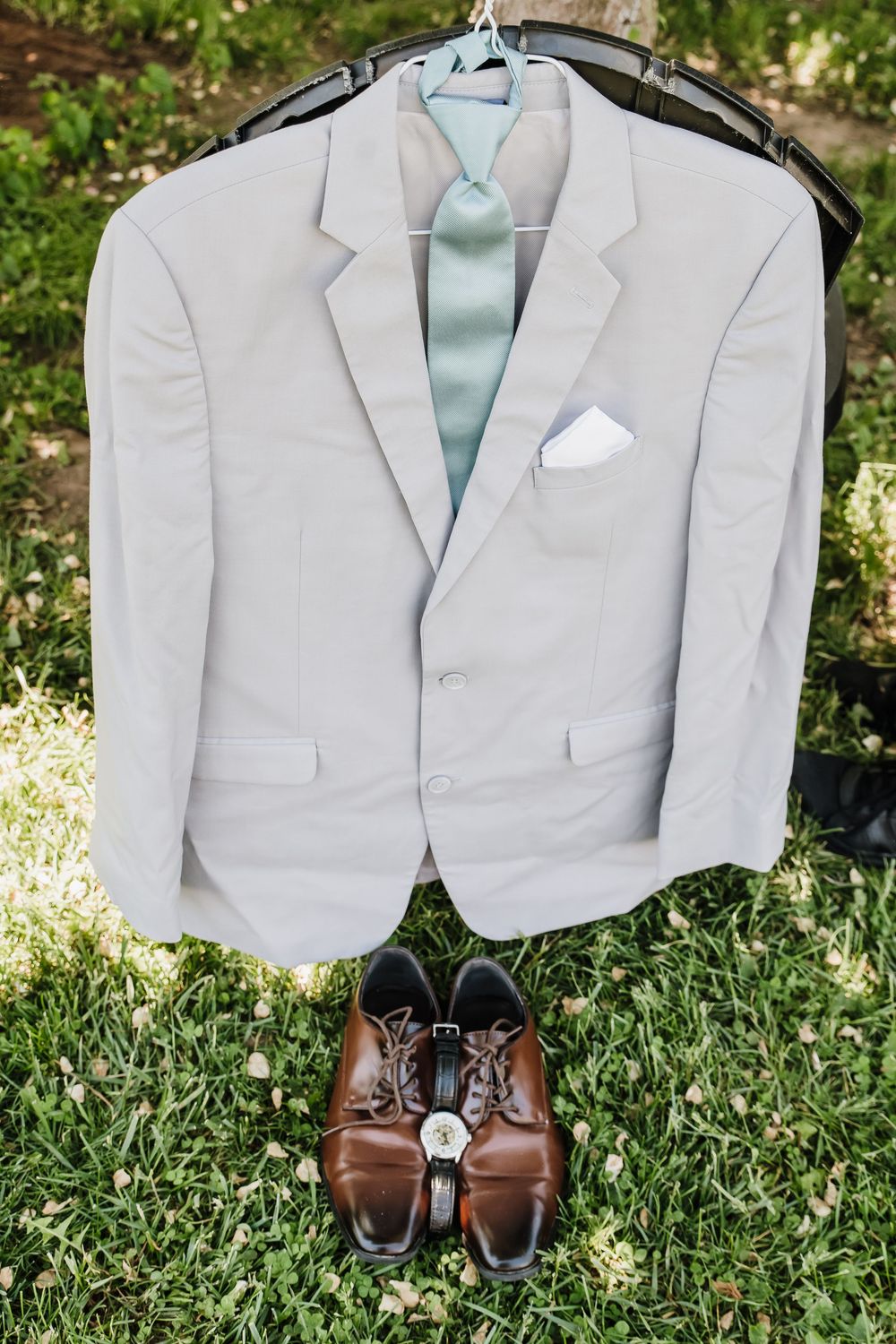 White suit jacket with mint tie on hanger displayed above brown dress shoes and watch on green grass.