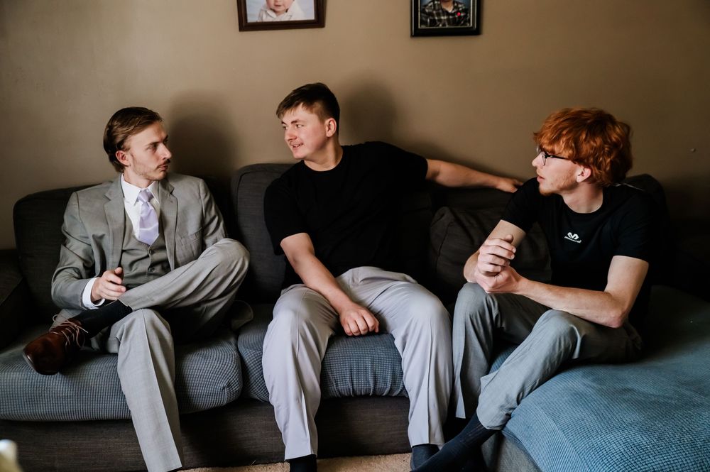 Three young men sitting on a gray couch having a casual conversation in a living room.