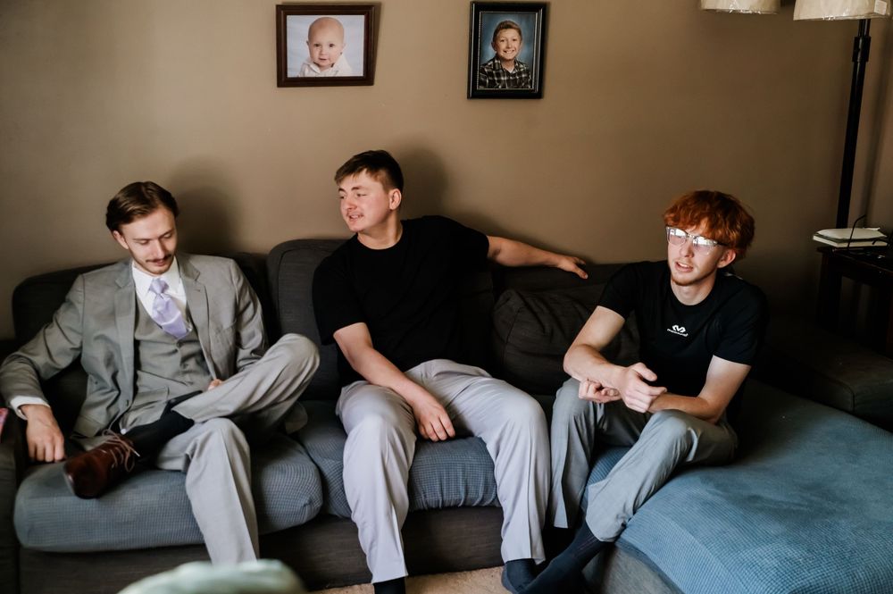 Three young men sitting on a couch in a living room with framed photos on the wall behind them.