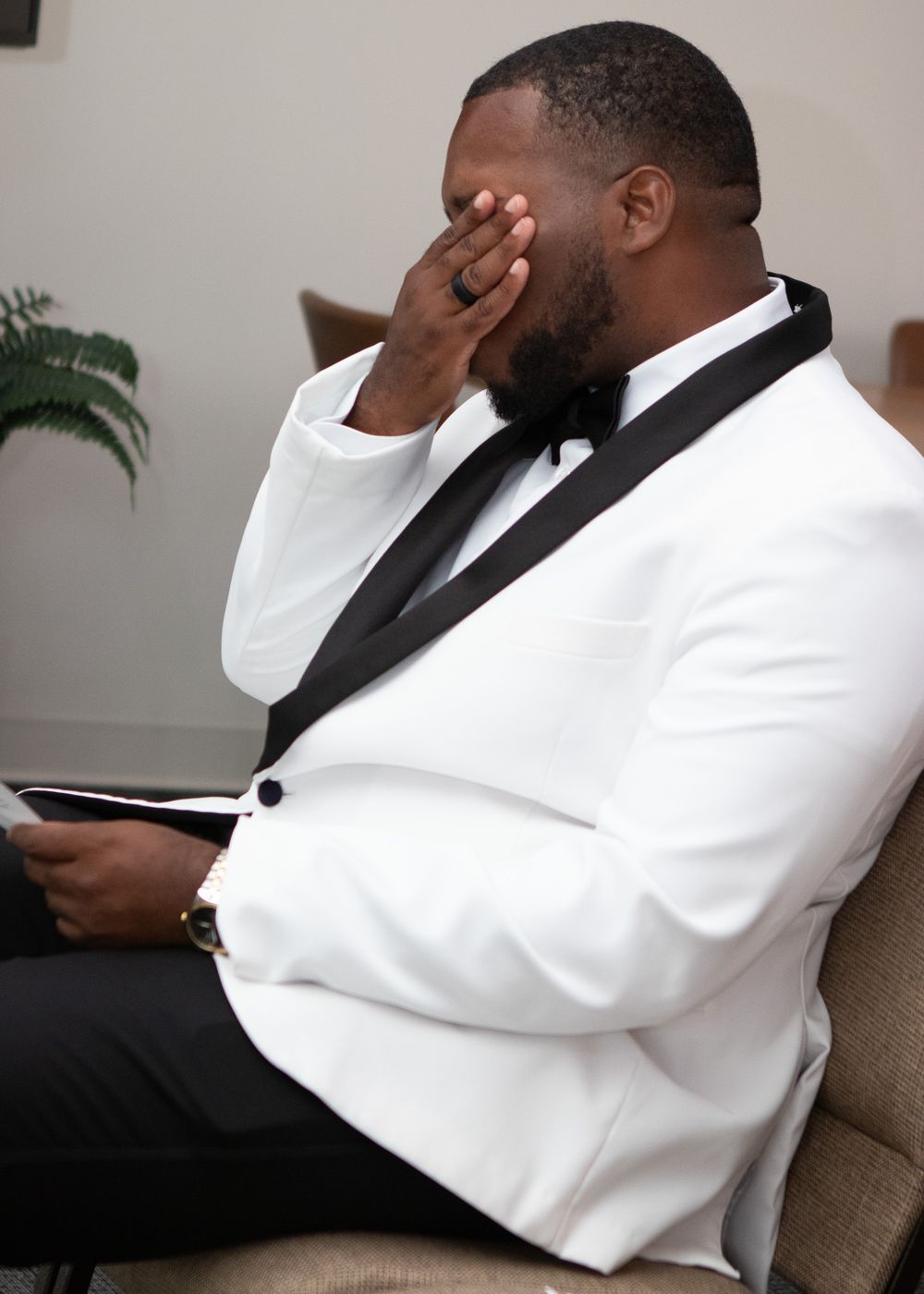 An emotional groom in a white tuxedo covers his face while reading wedding vows indoors.