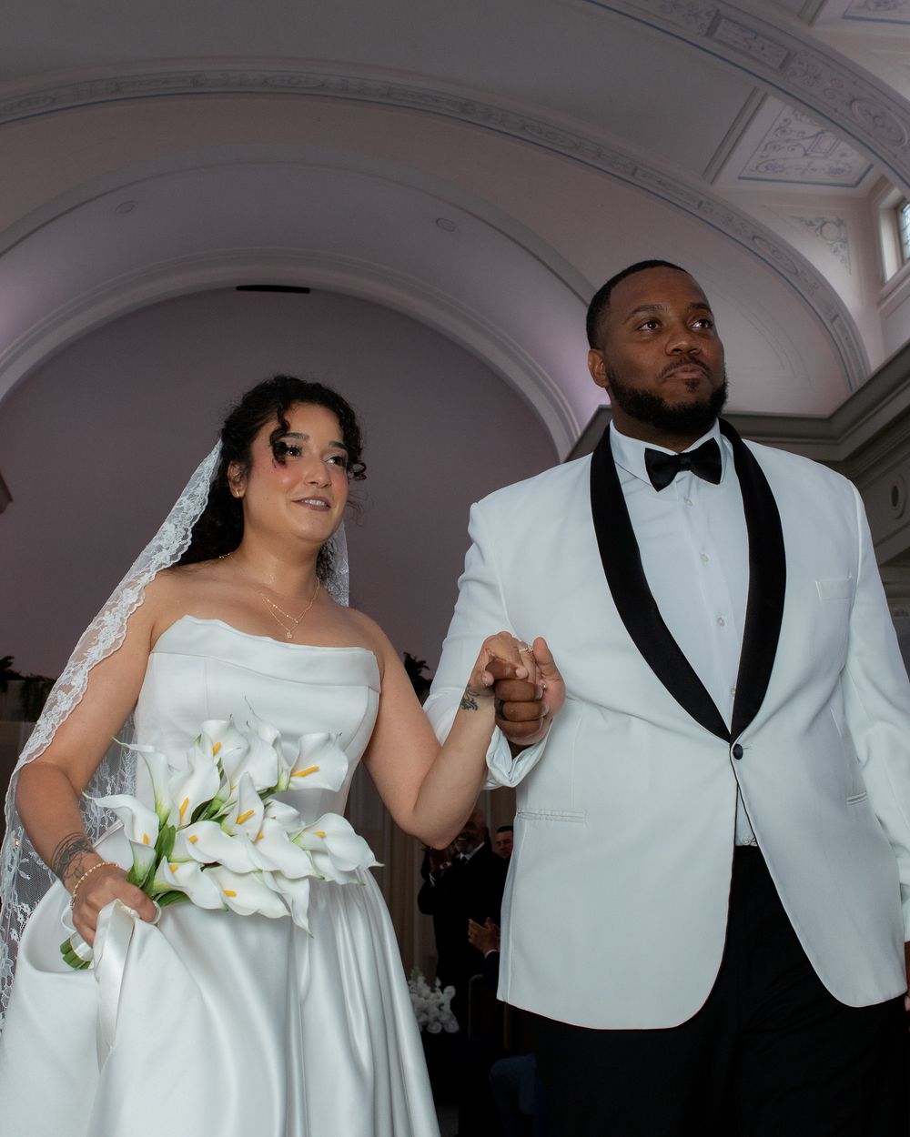 Newlyweds walk down church aisle, bride holding calla lily bouquet, groom in white tuxedo jacket.