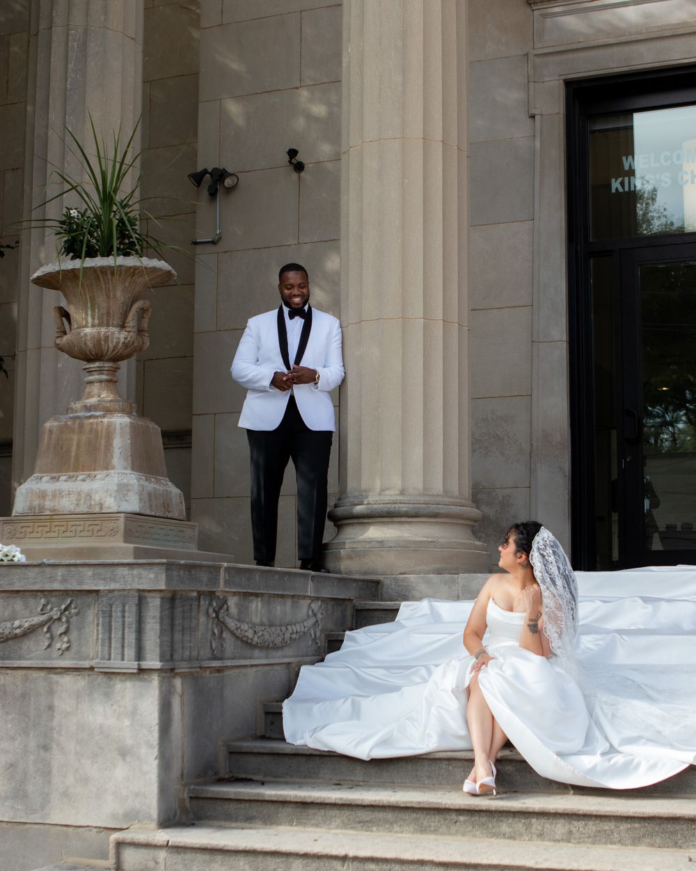 Groom in white tuxedo jacket stands on steps above bride in white wedding gown outdoors.