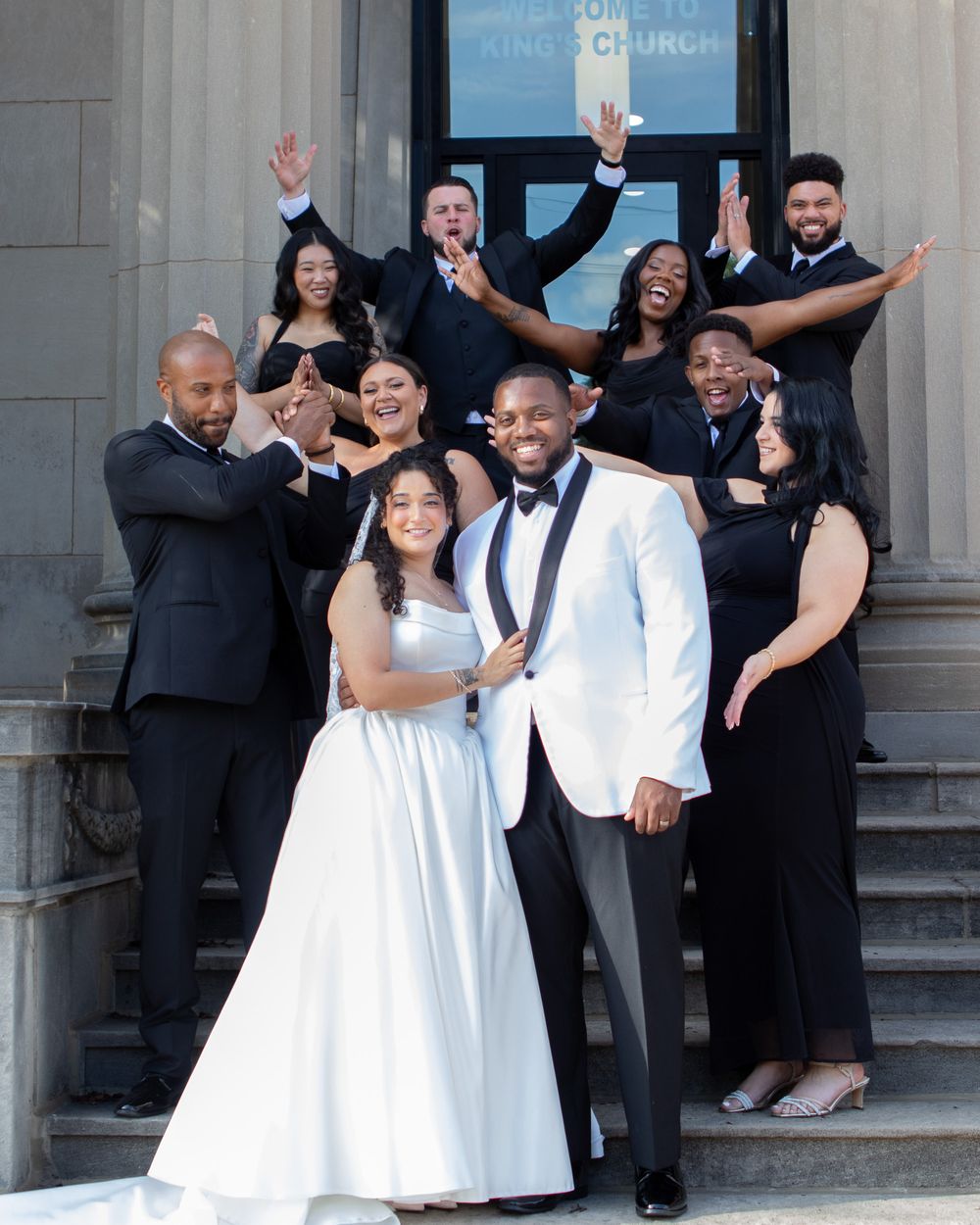 Wedding party in black attire celebrates with bride and groom on church steps.