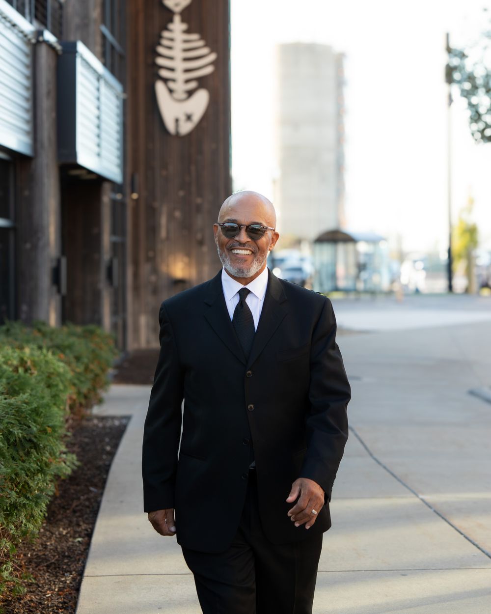 Elegant older man in black suit and sunglasses walks along sidewalk outside urban venue.