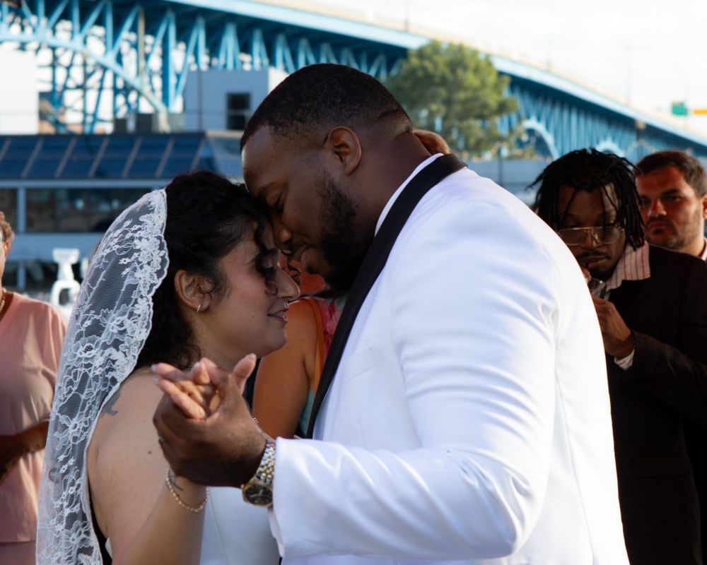 A bride in a white gown and veil shares an intimate moment with her groom in a white tuxedo at an outdoor wedding.
