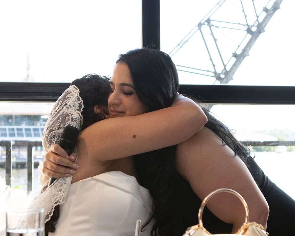 A bride in a white gown and lace veil receives a warm hug from a guest at her wedding reception.