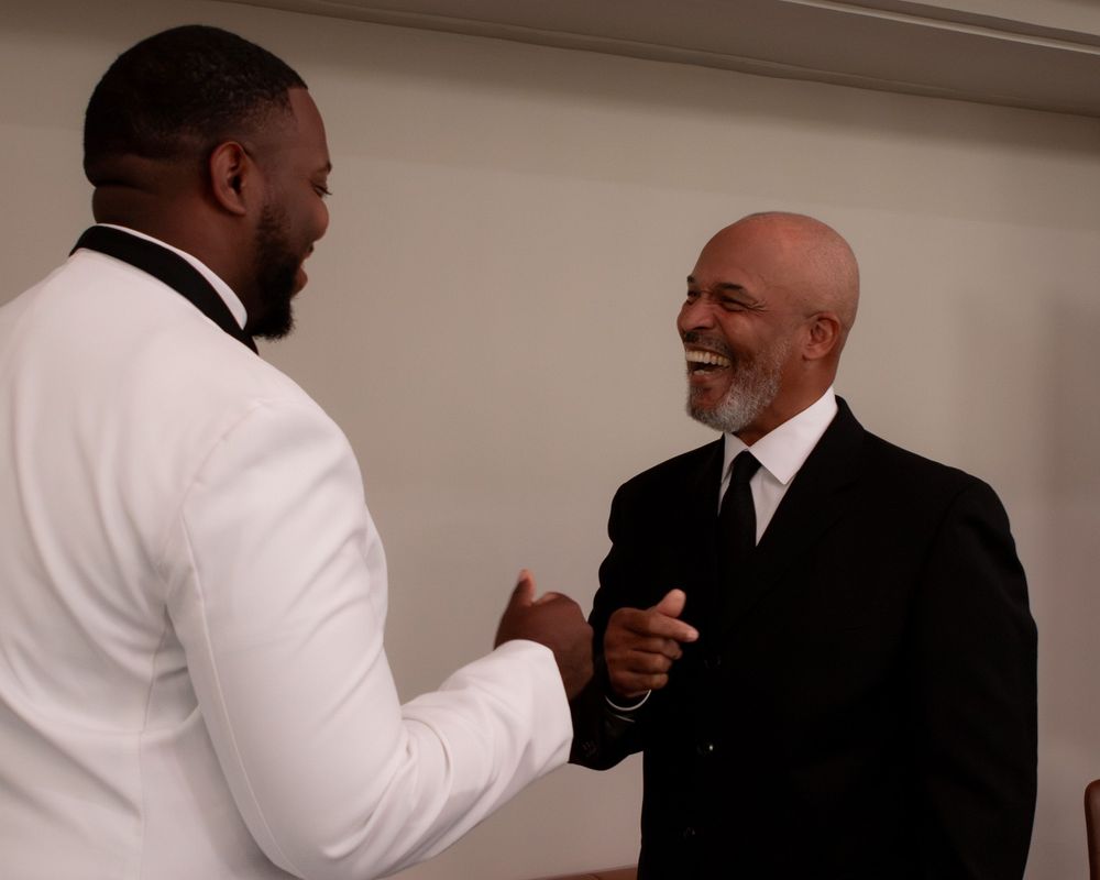 Groom in white tuxedo shaking hands and smiling with guest in black suit.
