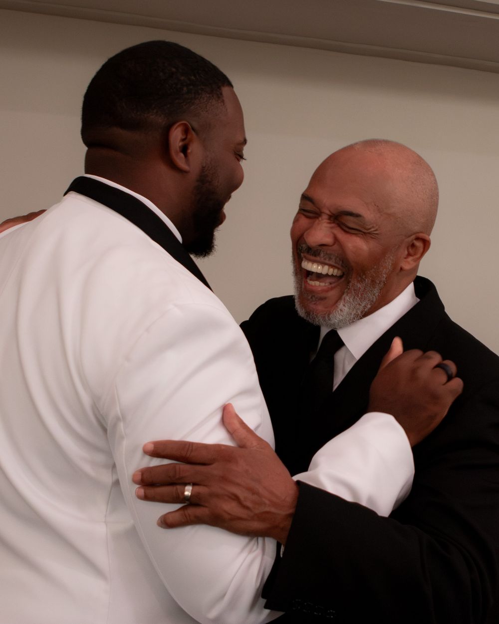 Groom in white tuxedo sharing warm embrace and laughter with older male wedding guest.