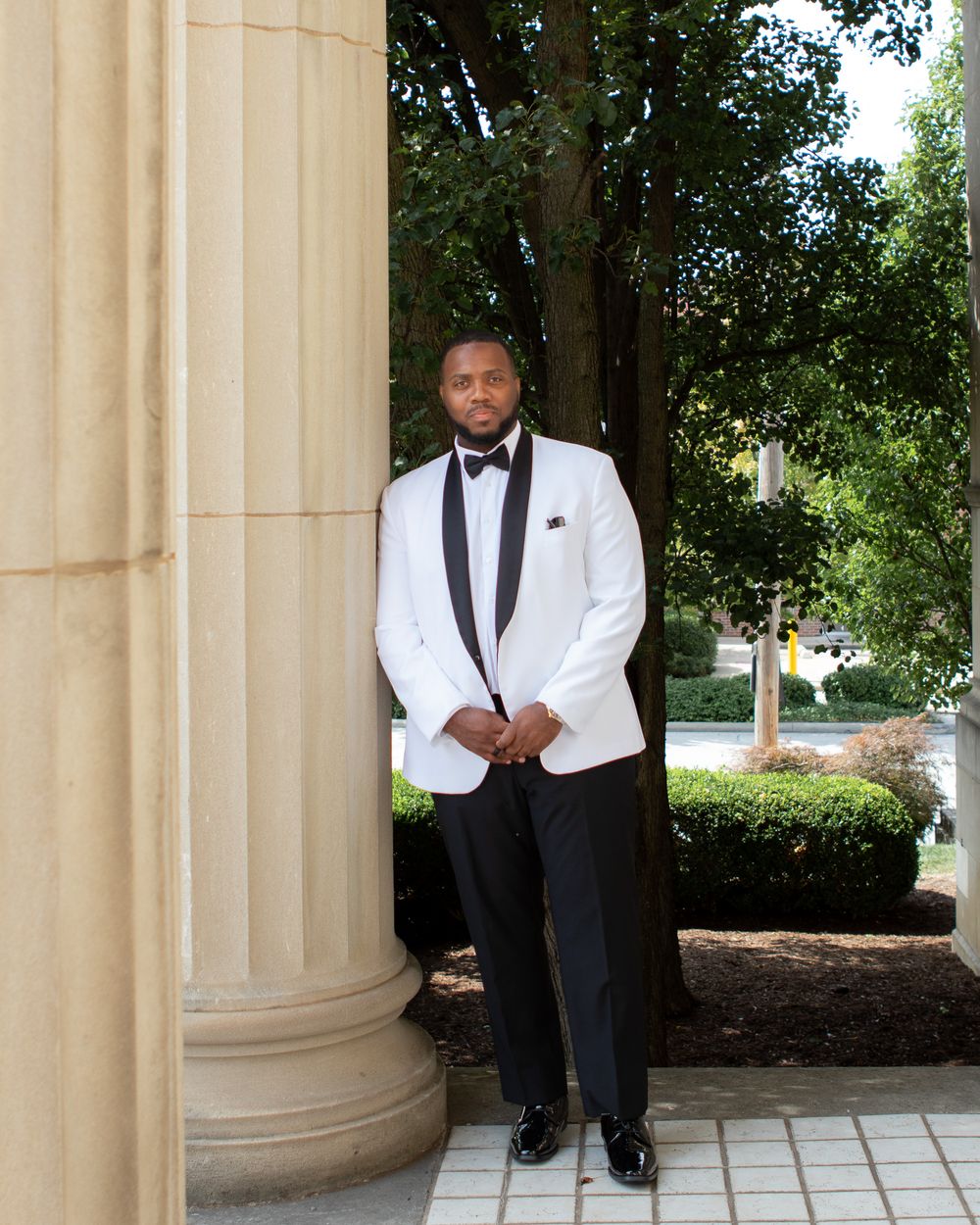 Groom in elegant white and black tuxedo posing beside stone column outdoors.