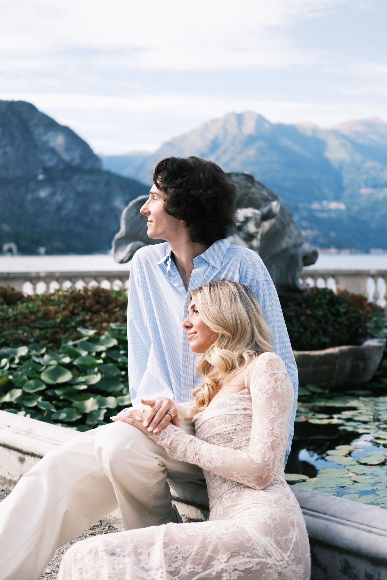 A couple sits by a lily pond with mountain views, the woman in lace dress leaning on a man in a blue shirt.