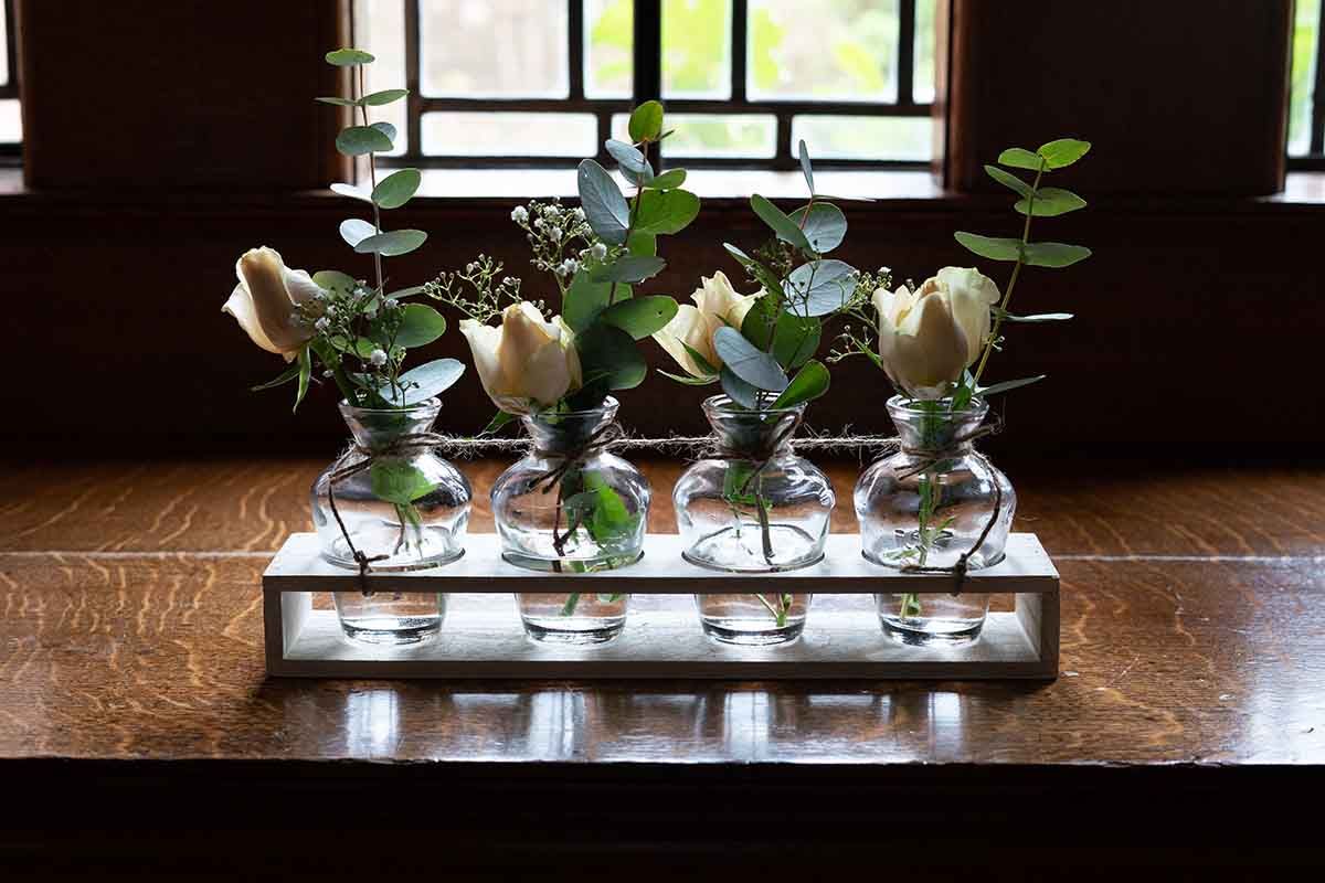 Five small glass bud vases holding white roses and greenery displayed on a white wooden tray on a wooden table.