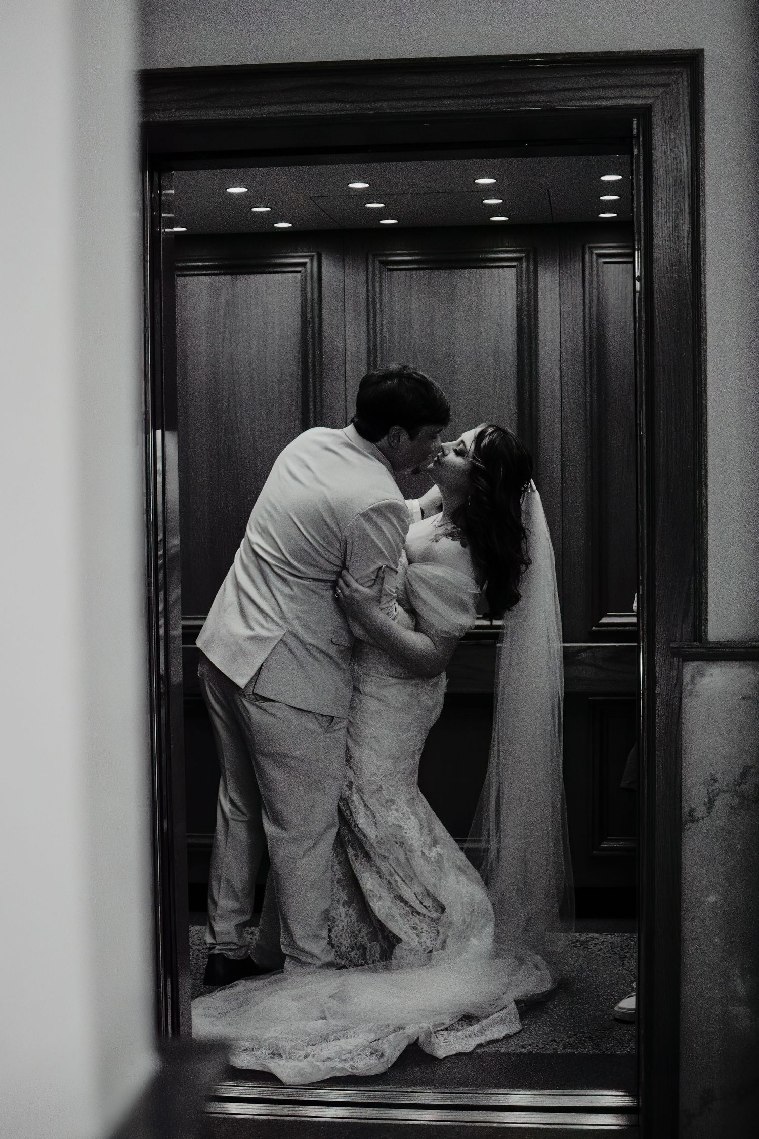 Black and white photo of a bride and groom sharing a romantic kiss inside an elegant elevator on their wedding day.