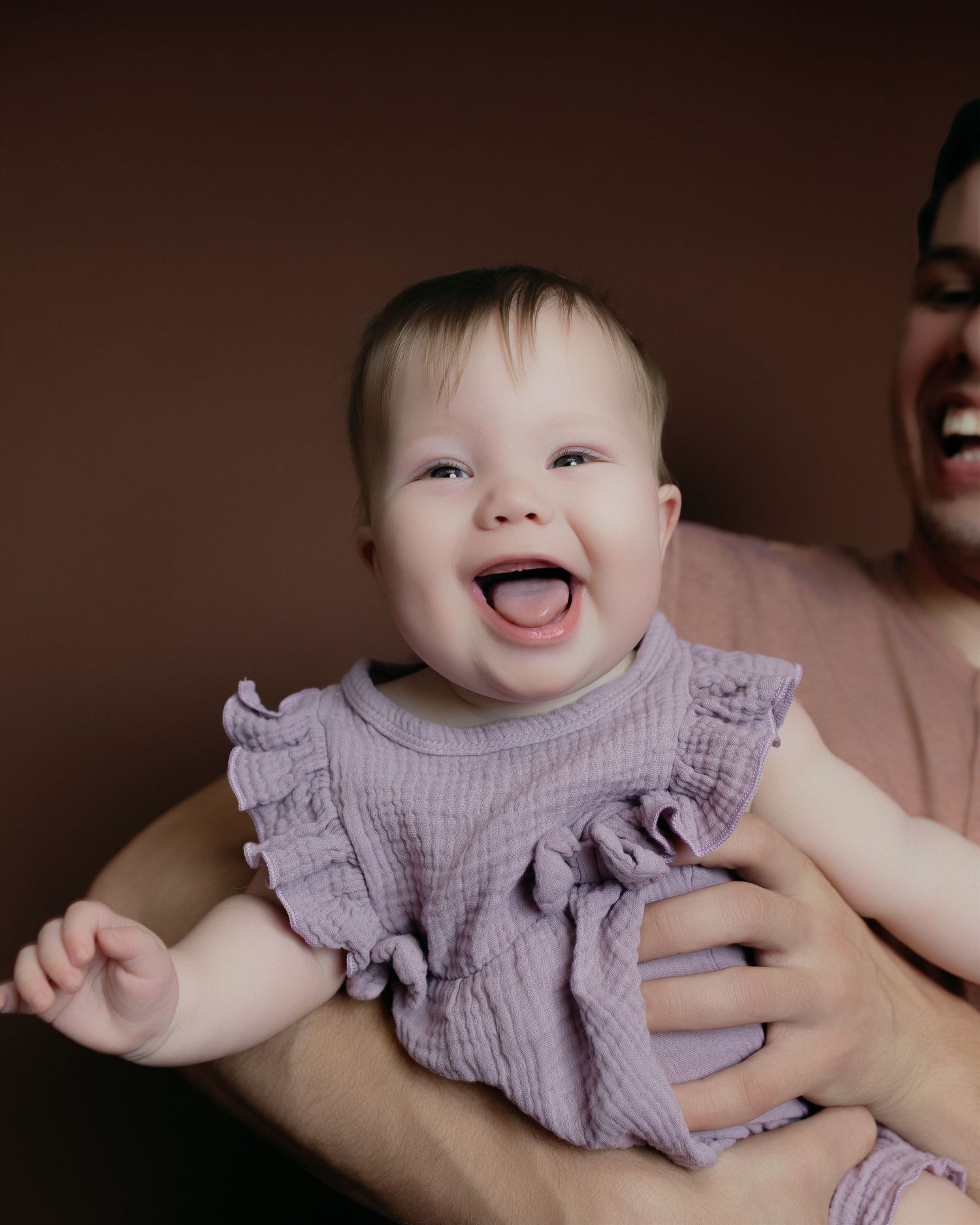 Happy baby in a lavender ruffled dress smiles joyfully while being held, photographed against a dark brown background.