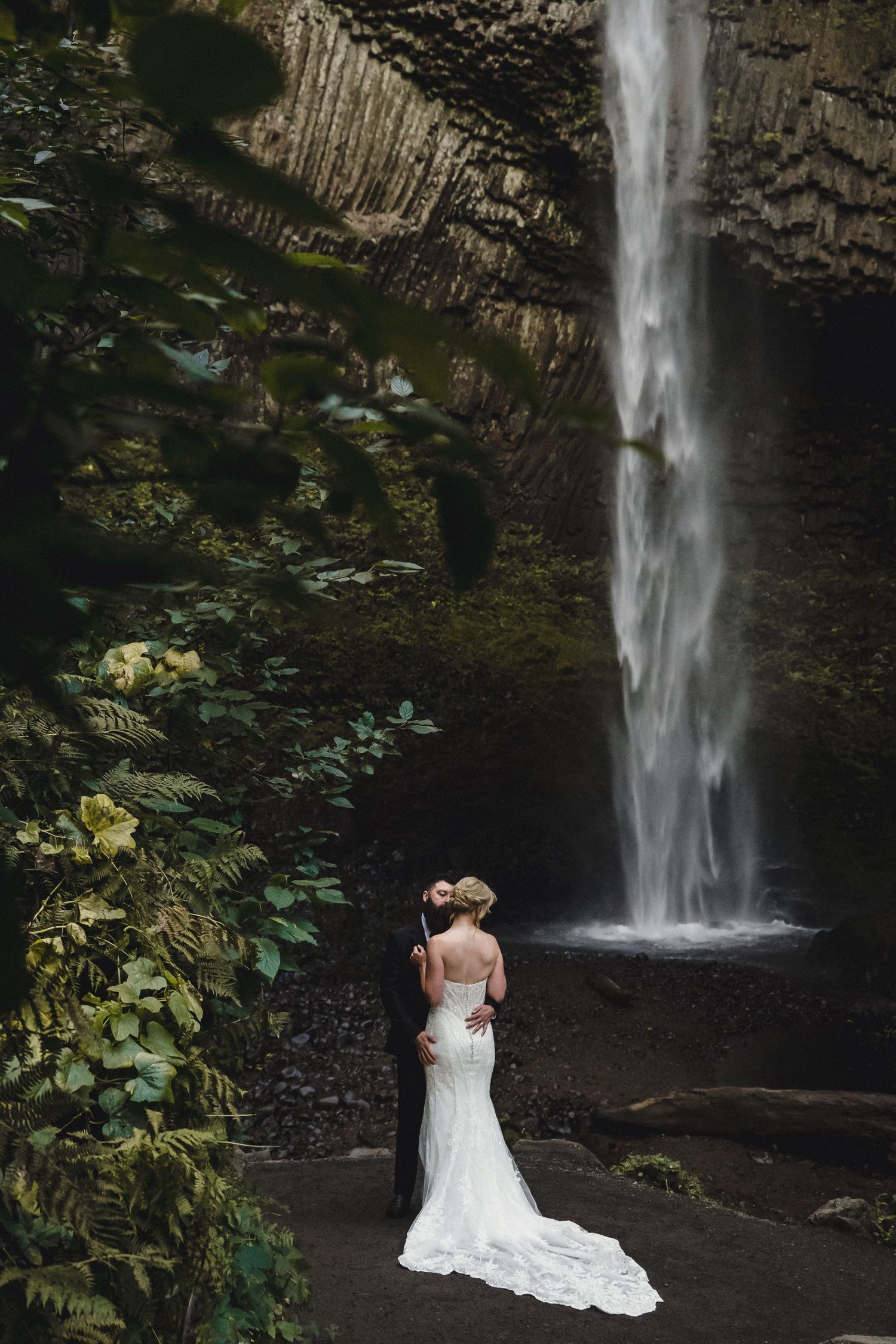 Bride and groom embrace before a stunning waterfall surrounded by lush greenery in a romantic outdoor wedding setting.