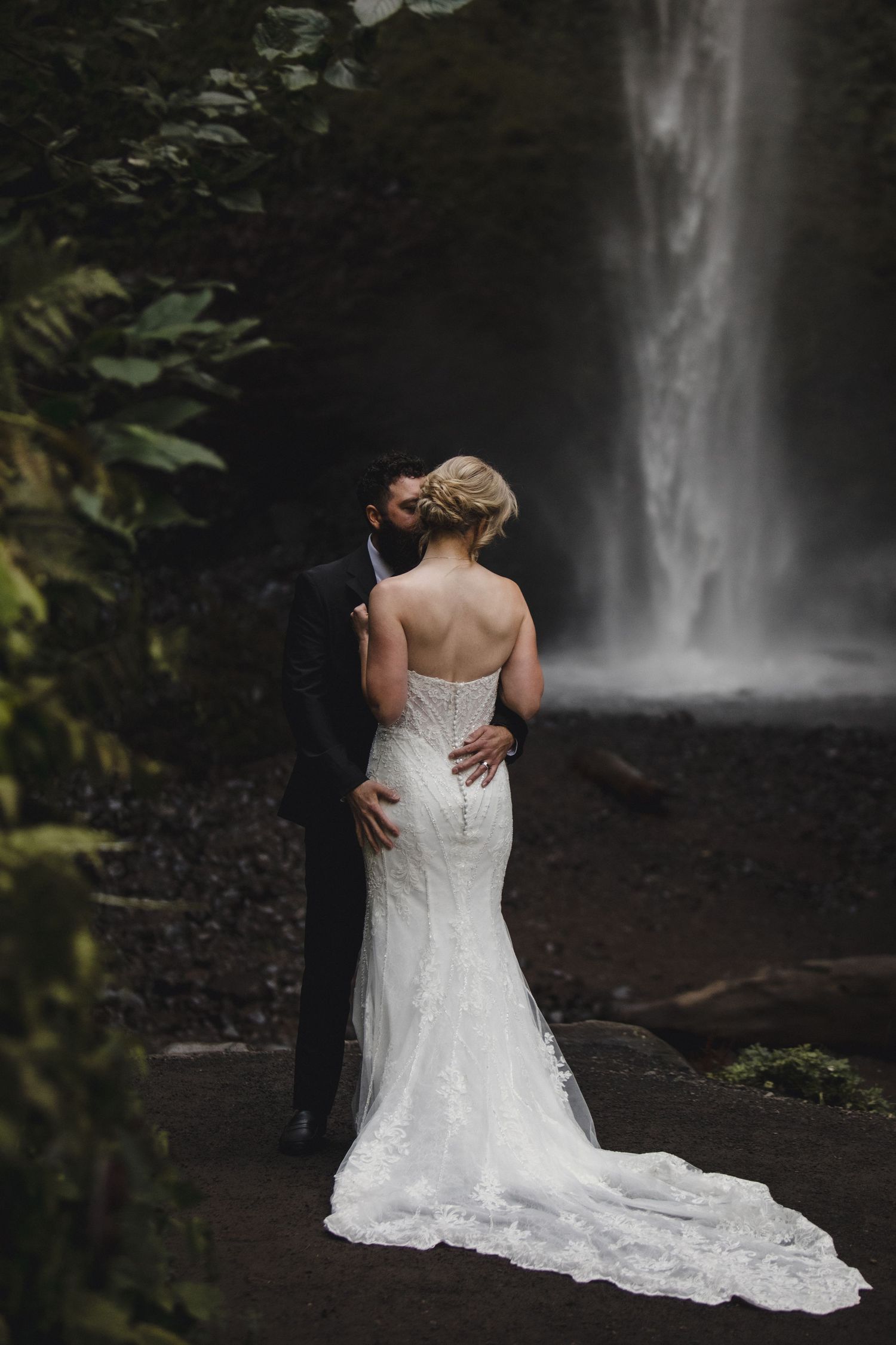 Bride and groom embrace before a misty waterfall, her lace mermaid gown trailing on dark stones.