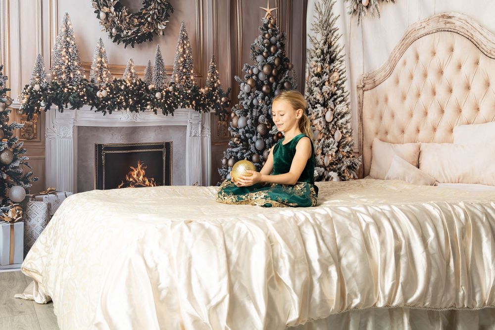 Girl in green dress sits on bed holding gold ornament near decorated Christmas trees and lit fireplace.