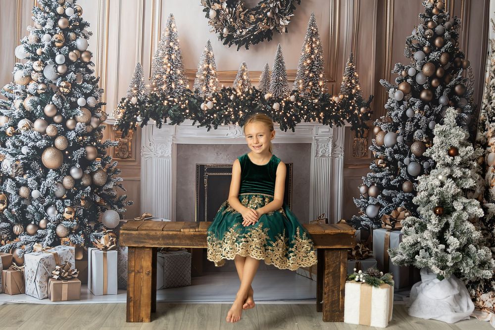 Girl in green gold dress sits on bench surrounded by Christmas trees and fireplace in elegant holiday setting.
