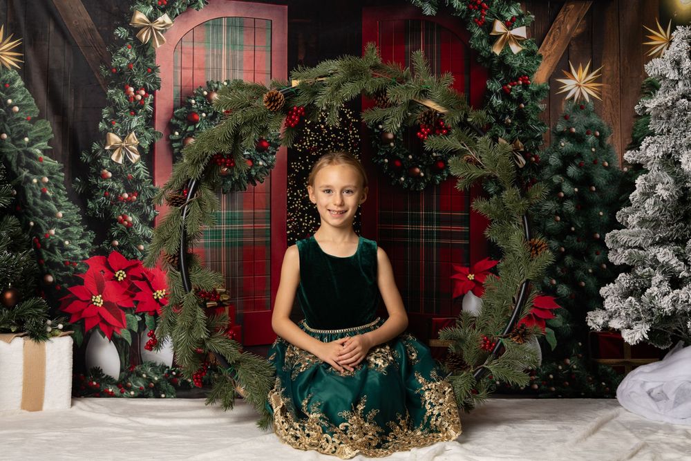 Girl in green velvet dress poses inside large Christmas wreath with holiday backdrop and decorated trees.