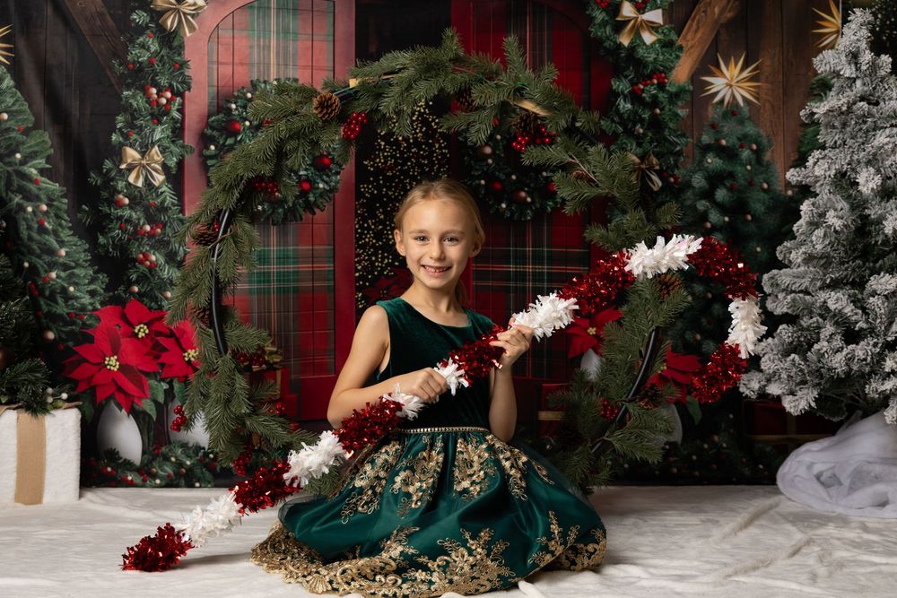 Young girl in green gold dress holds red white garland among Christmas trees and festive holiday decorations.