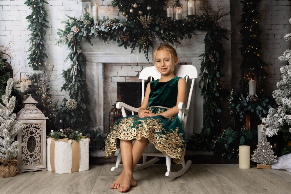 Girl in green velvet dress sits in rocking chair by decorated Christmas fireplace with garland and candles.