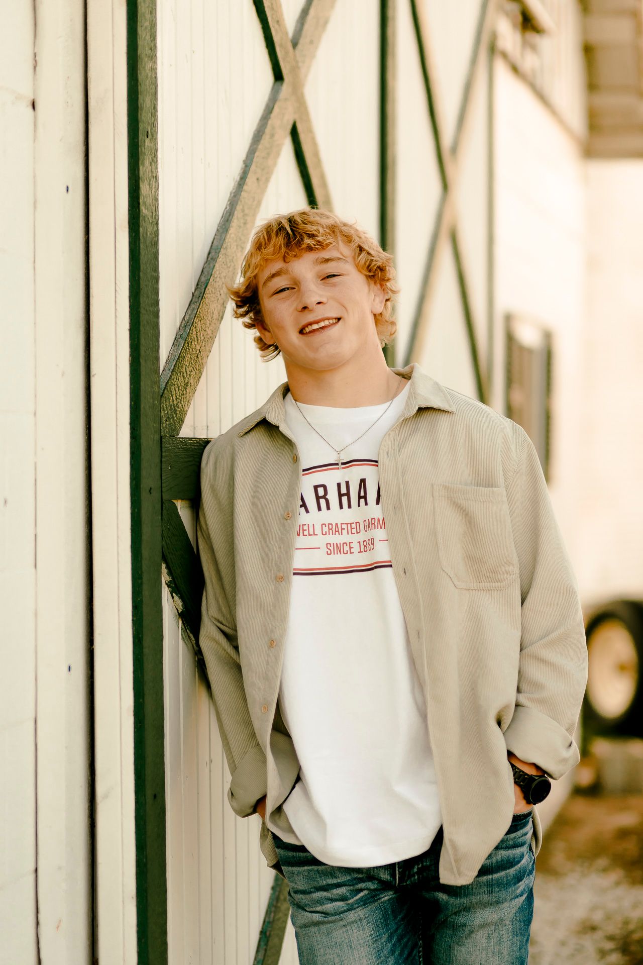 Smiling teenage boy with curly blonde hair wearing a Carhartt t-shirt and gray jacket, leaning against a white barn wall.