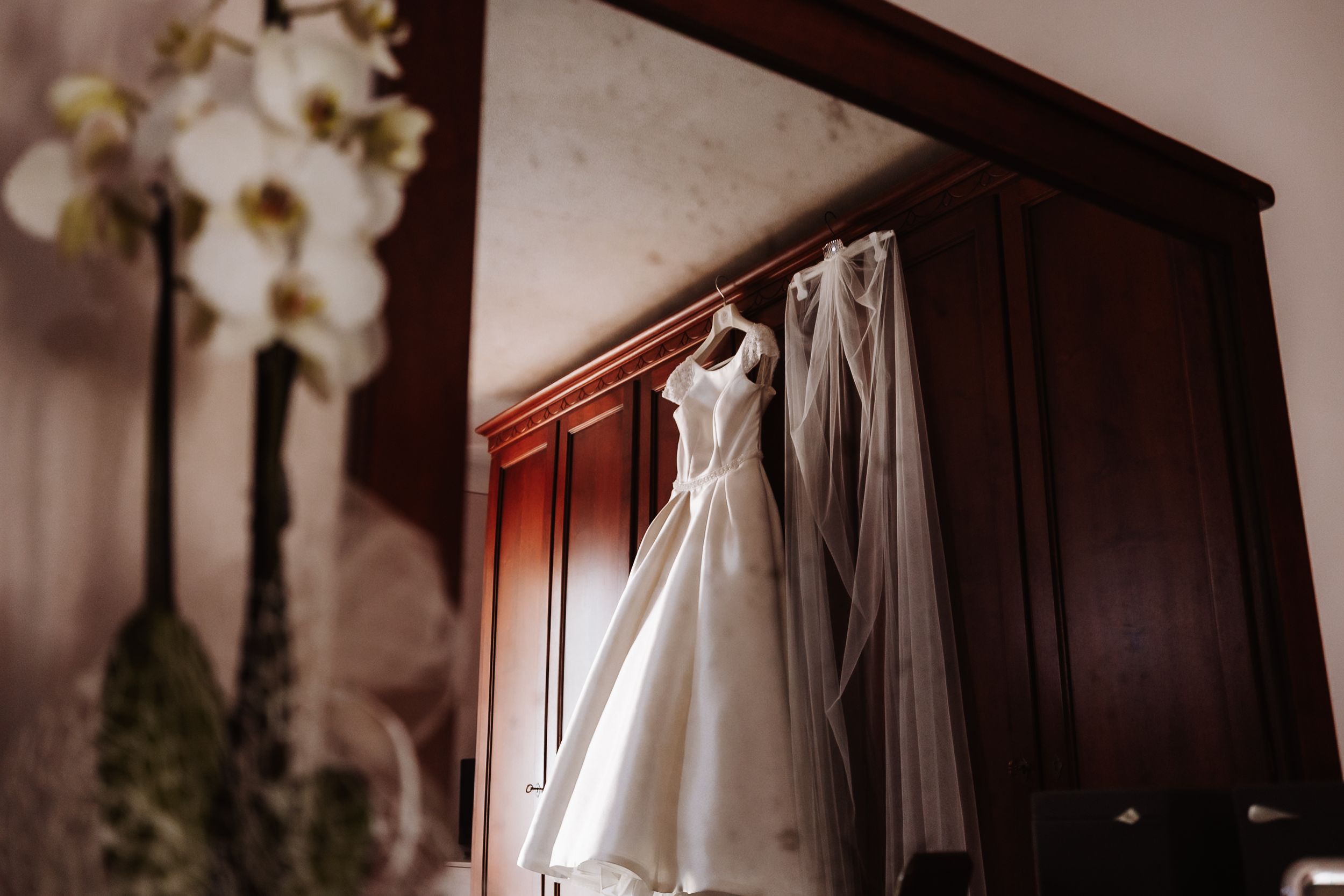Elegant white wedding dress with veil hanging on a dark wooden wardrobe beside white orchid flowers.