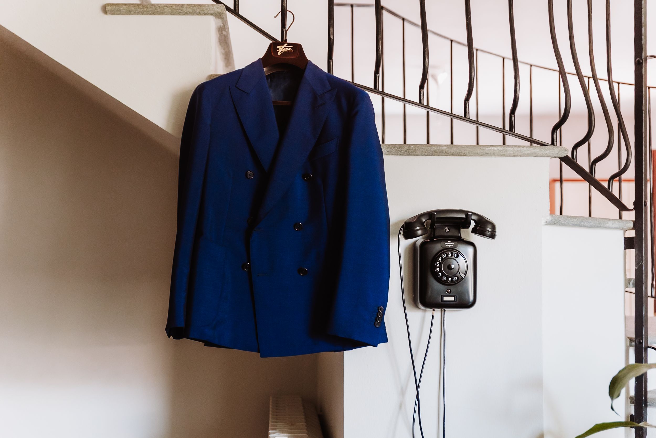 A navy blue double-breasted suit jacket hanging on a hanger near a vintage black rotary wall telephone.