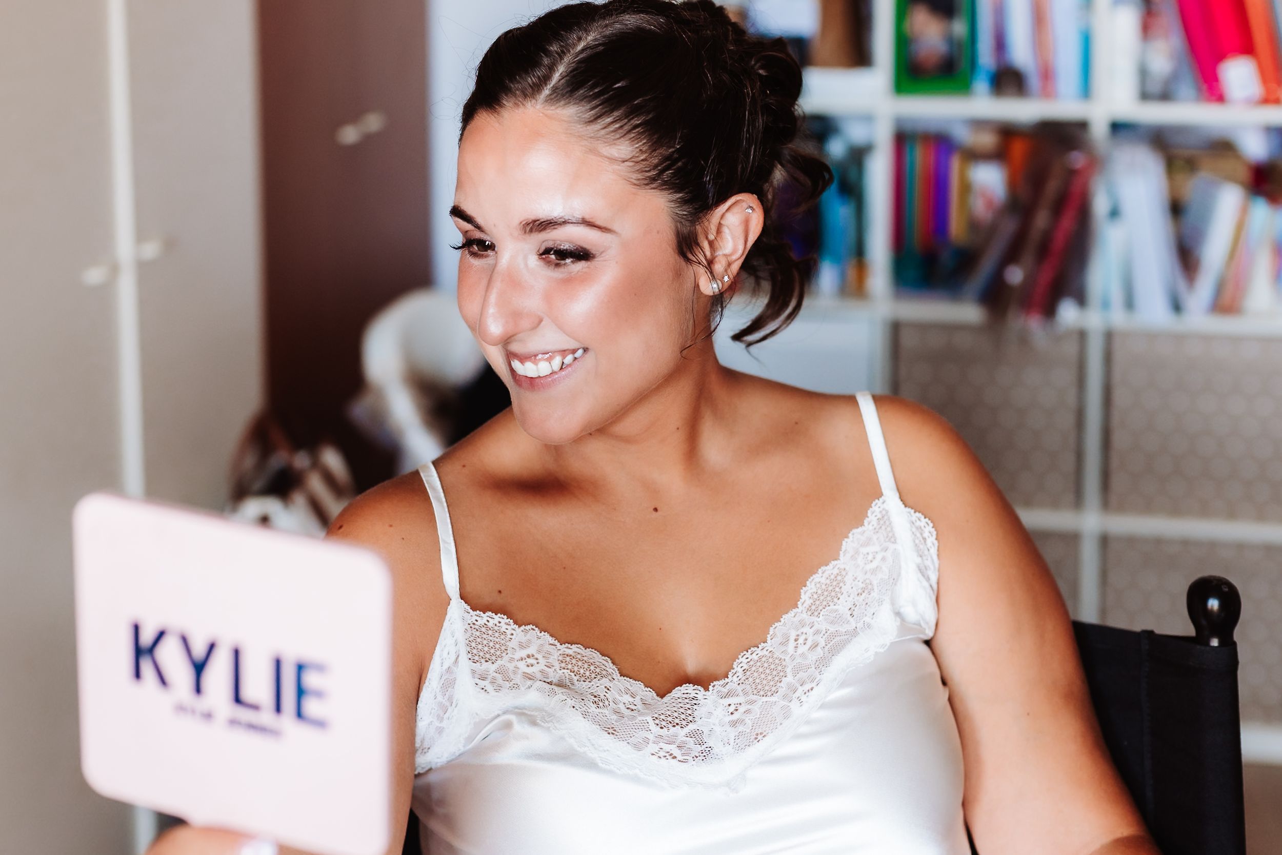 Woman smiling while holding a Kylie Cosmetics mirror, wearing a white lace top in a bright room.