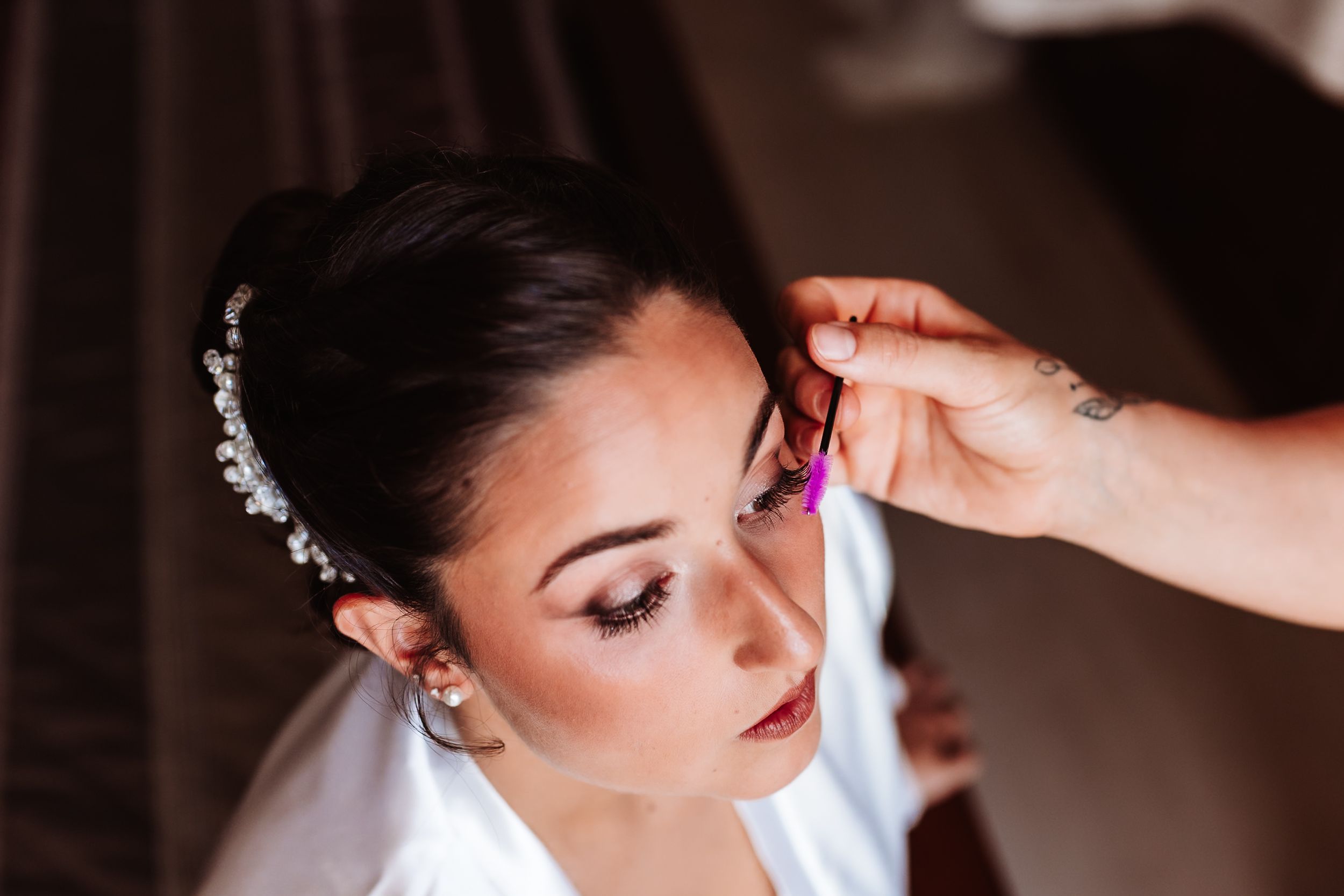 Bride in white robe getting earrings put on, wearing pearl headband and elegant makeup on her wedding day.