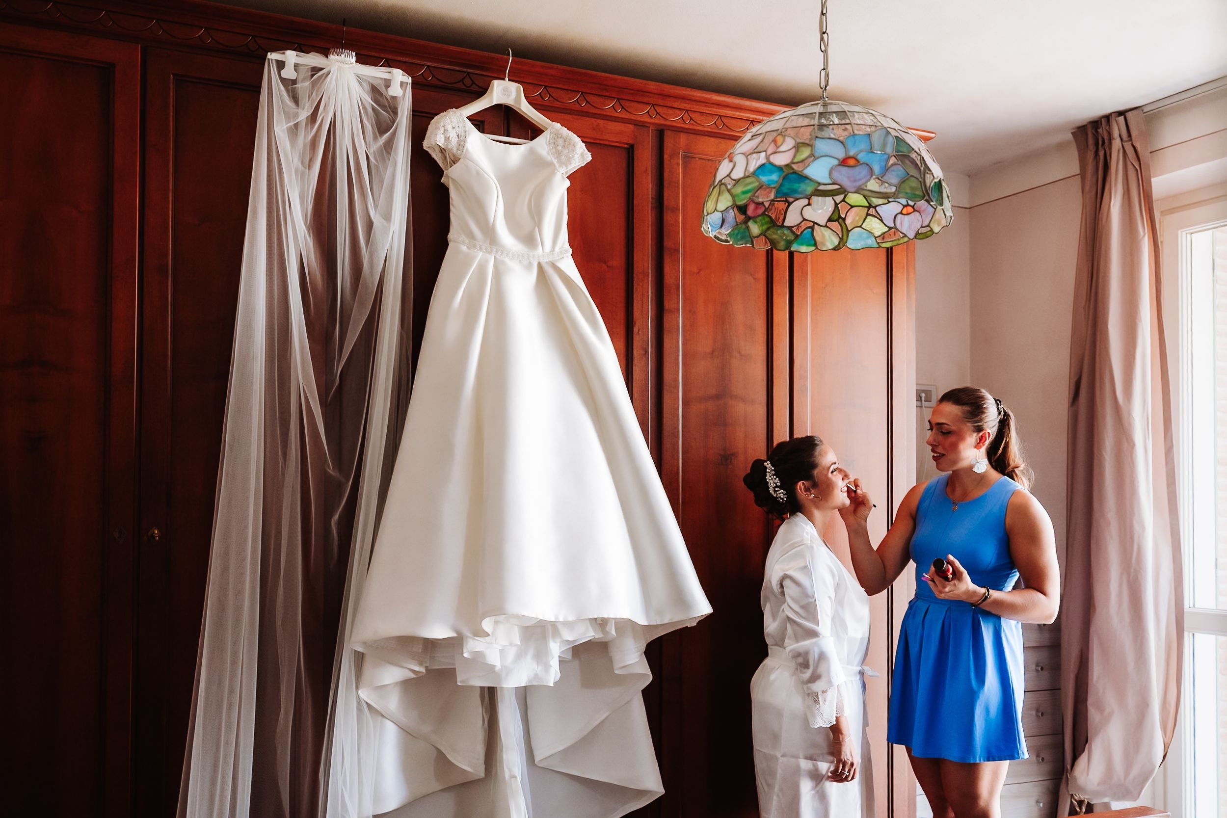 Bride in white robe gets makeup done by bridesmaid in blue dress, with elegant wedding gown hanging nearby.