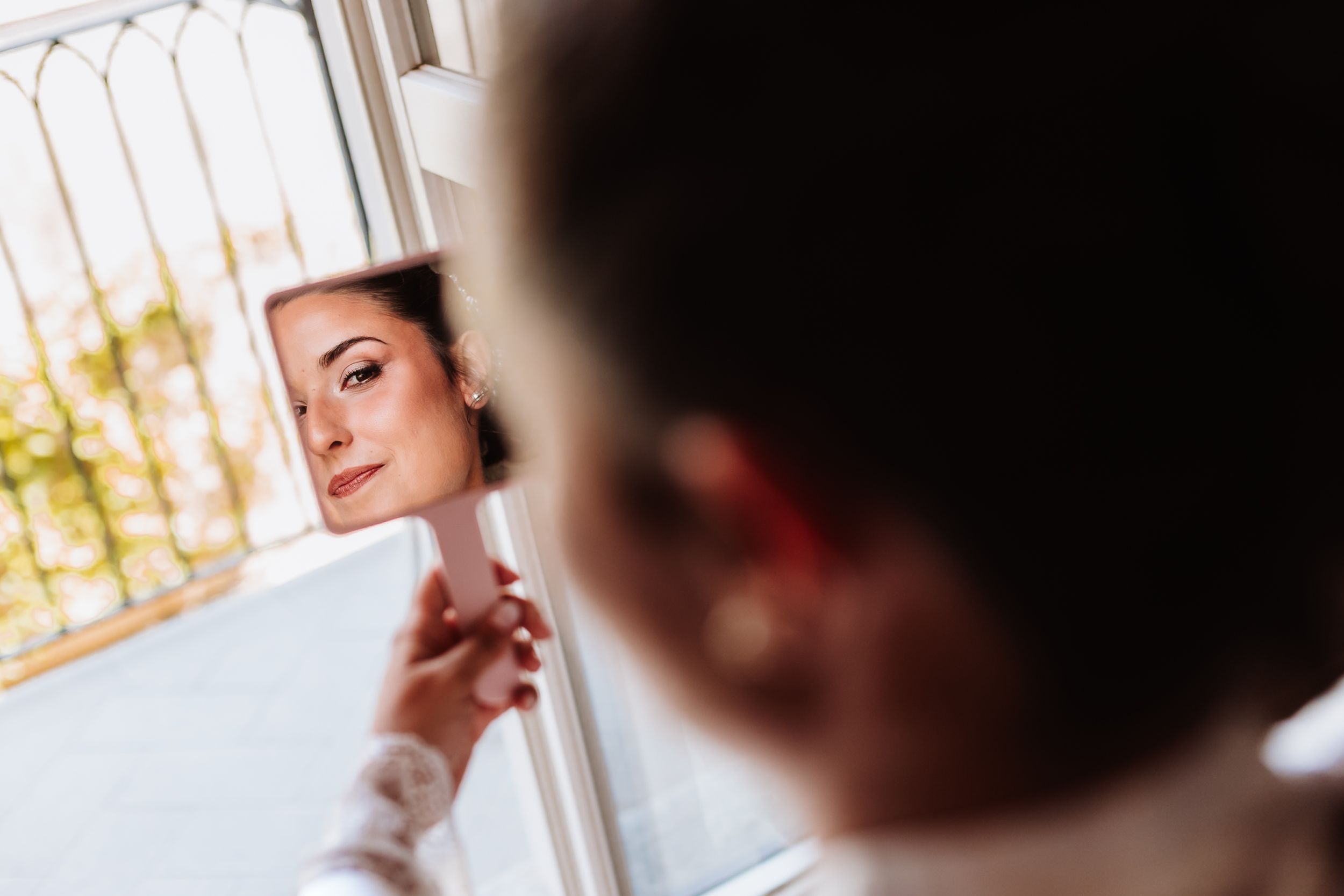 Bride gazes at her reflection in a handheld mirror near a window, capturing a serene pre-wedding preparation moment.