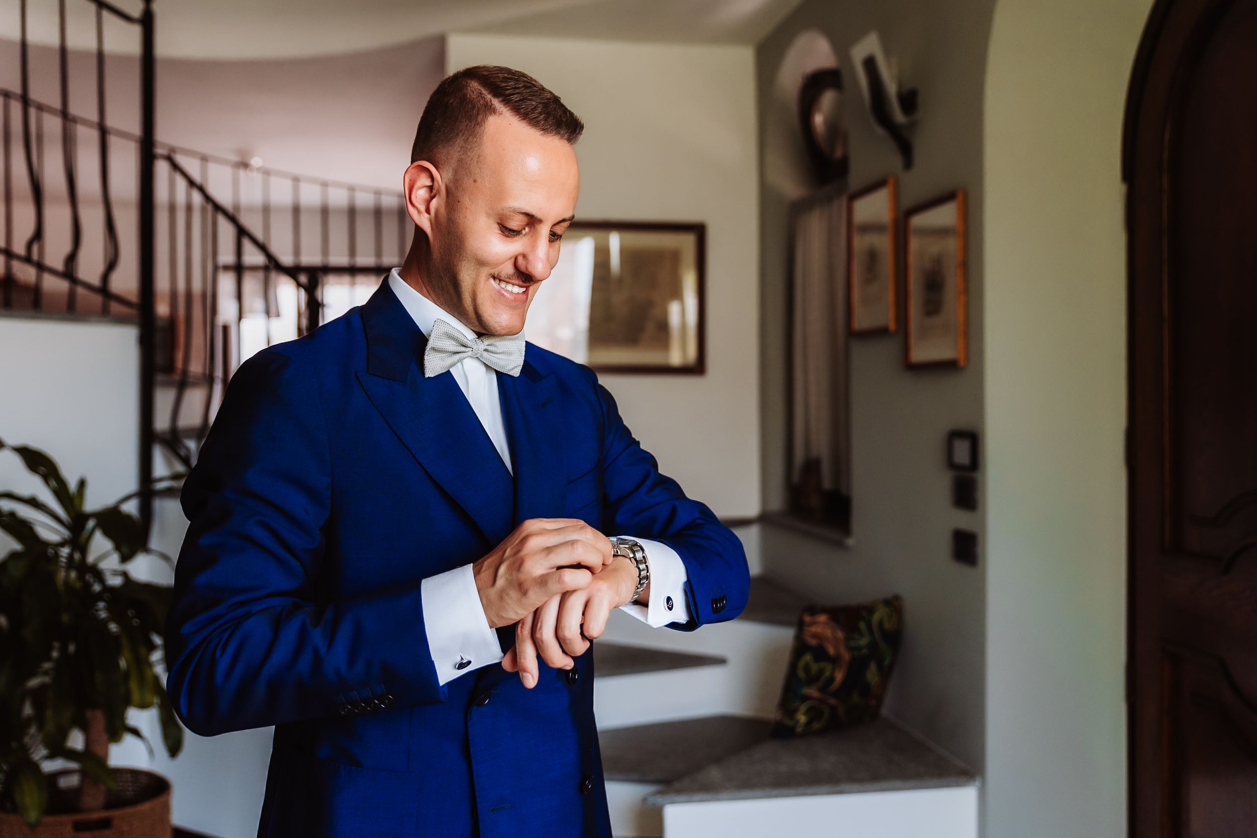 Elegant groom in blue suit and bow tie checking his watch while smiling before wedding ceremony.