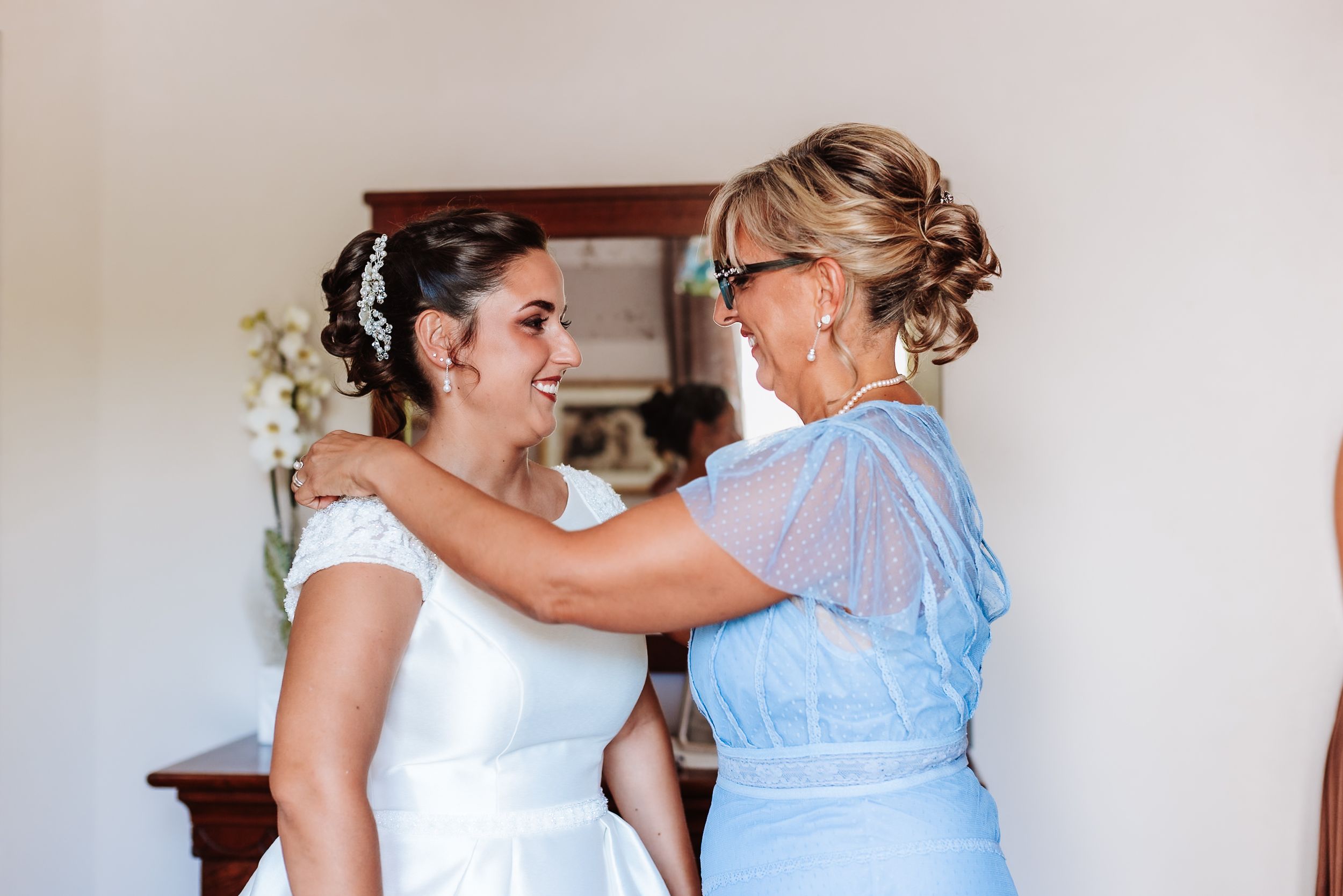 Bride in white dress smiling with woman in blue dress adjusting her shoulders indoors.
