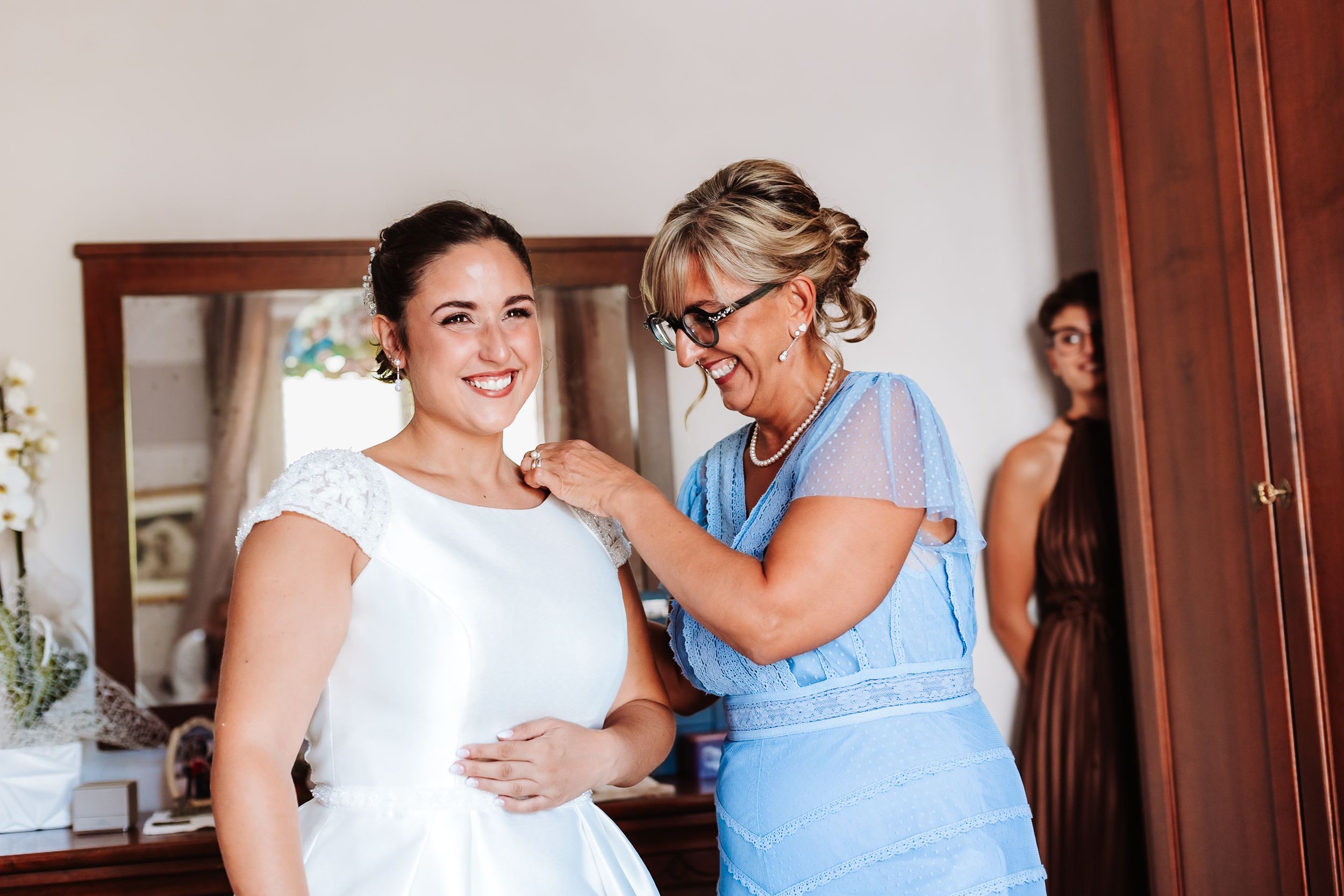 A woman in a white wedding dress smiles as an older woman in blue helps her prepare on her wedding day.
