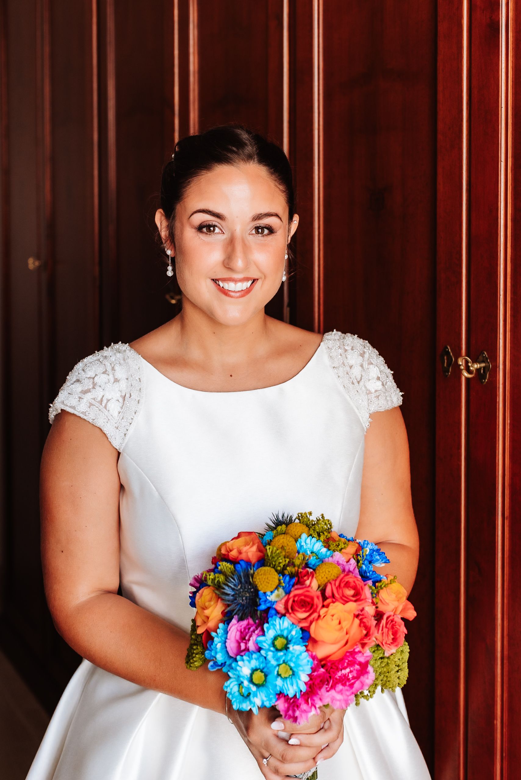 Smiling bride in white lace dress holding vibrant colorful bouquet of pink, blue, and orange flowers.