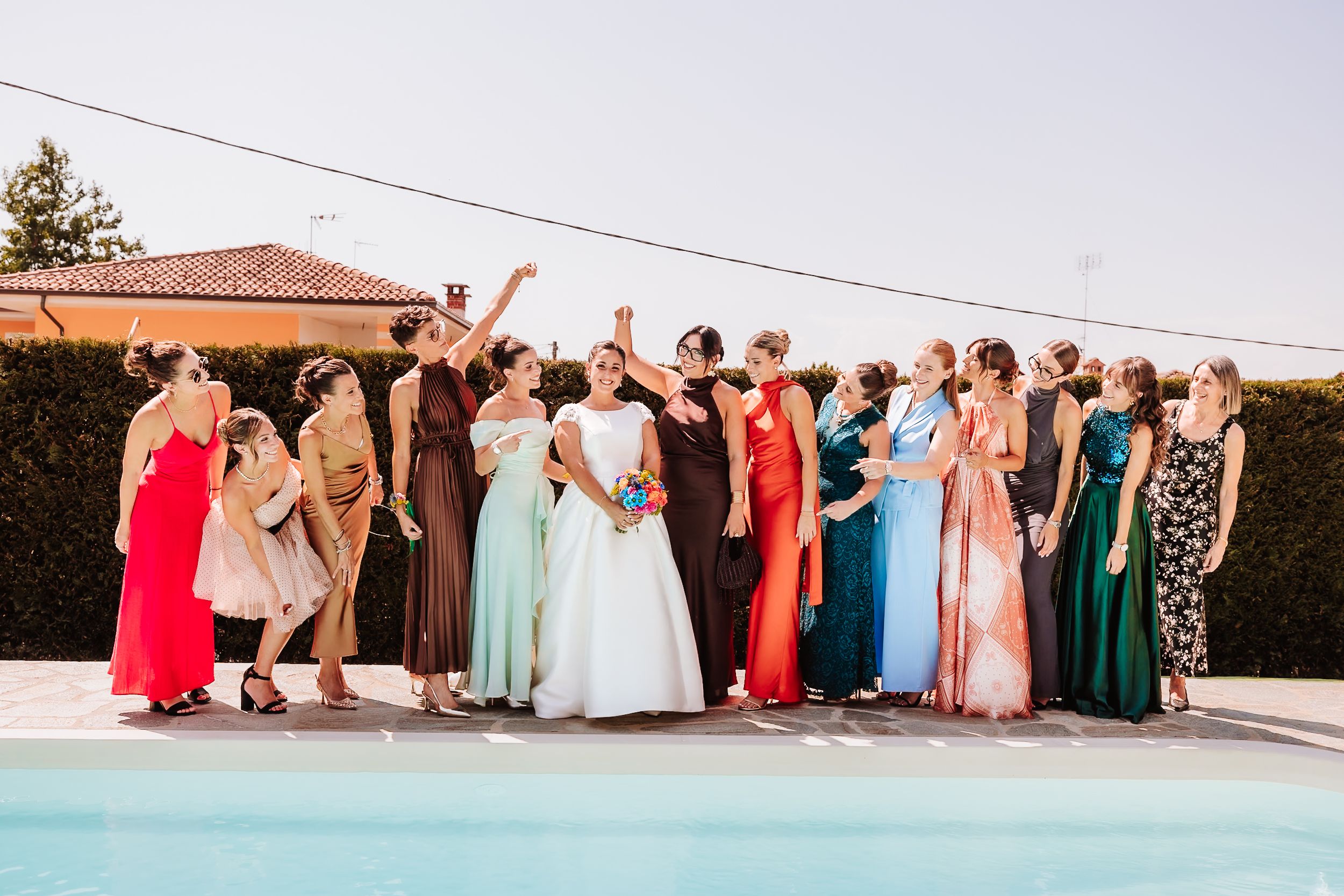 Bride in white gown surrounded by colorful bridesmaids celebrating poolside at an outdoor wedding reception.