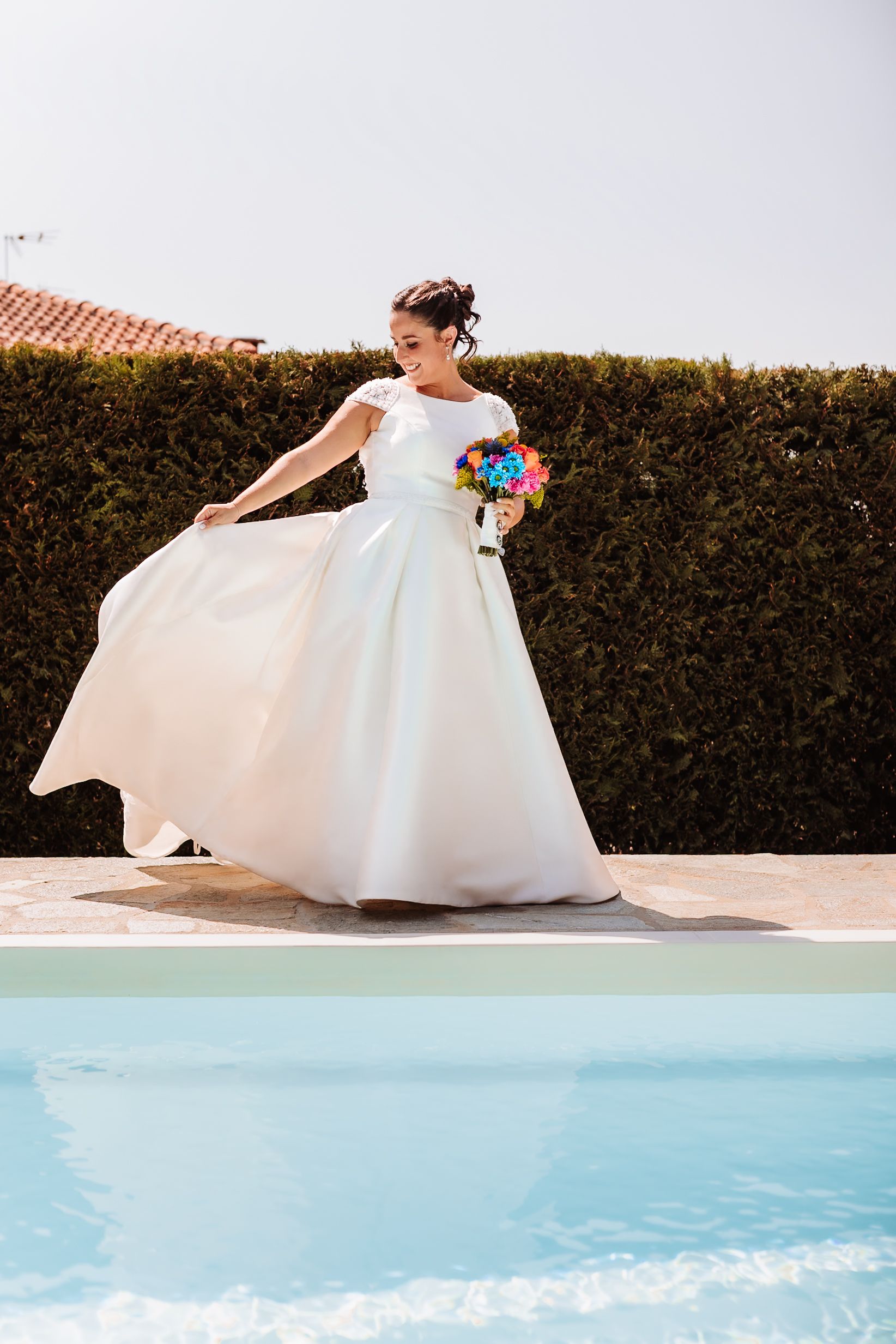 Bride in white gown twirling with colorful bouquet beside a pool, hedge backdrop on sunny day.
