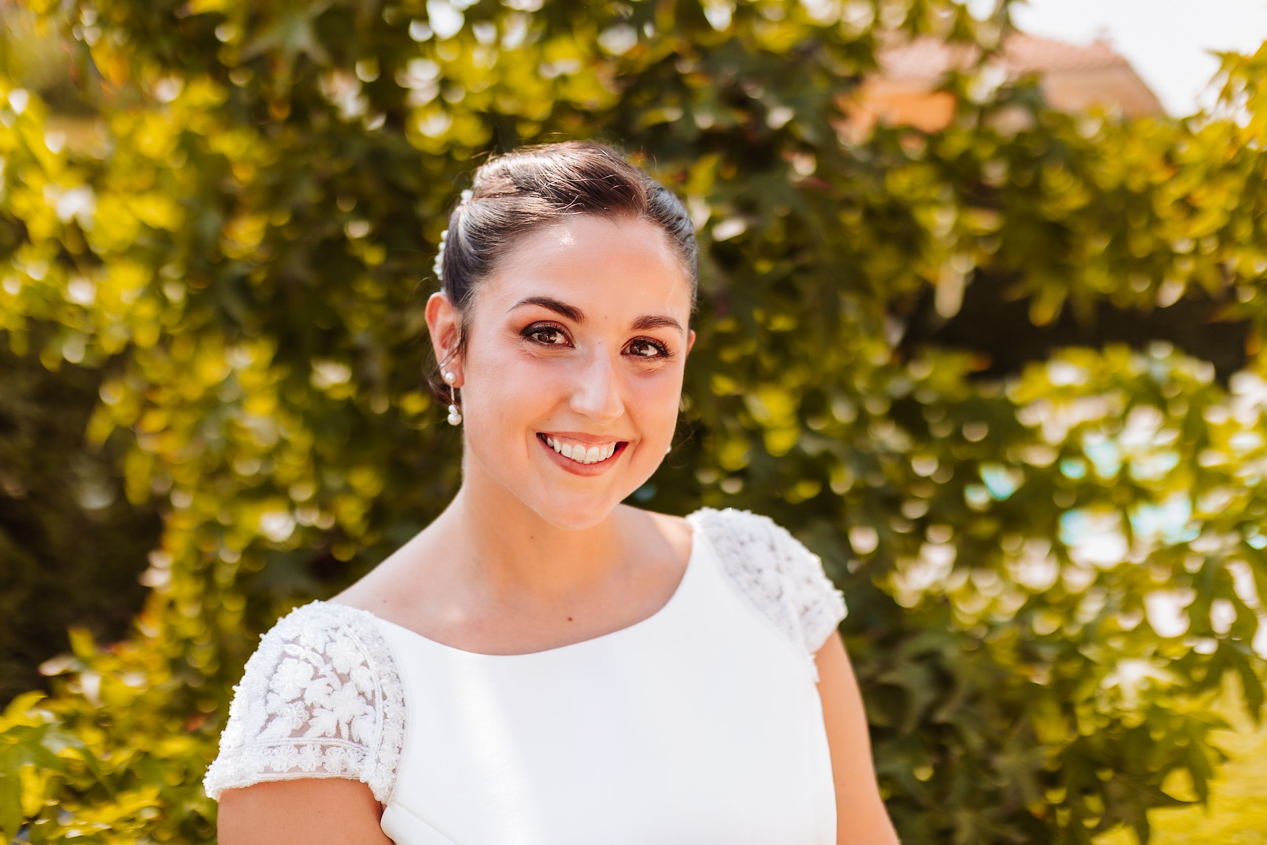 Smiling bride in white lace dress with updo hairstyle posing outdoors with green trees in background.